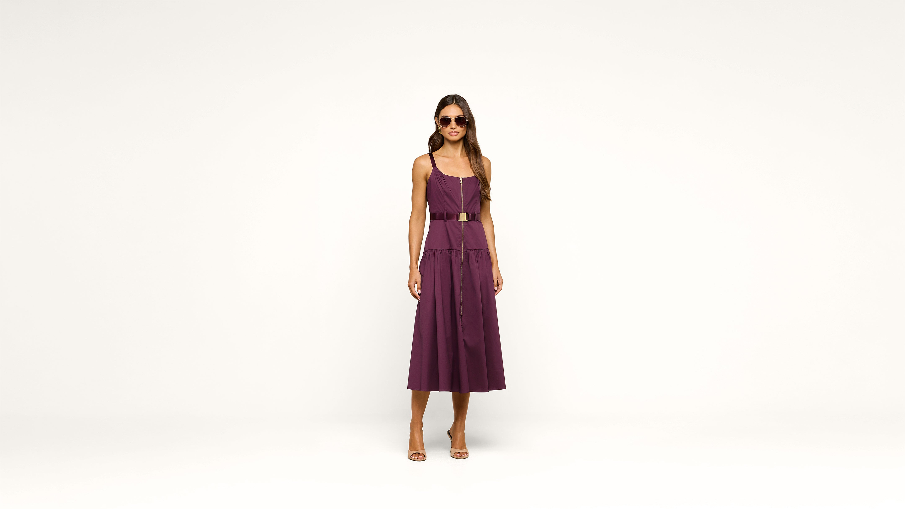 A woman wearing a sleeveless purple dress, sunglasses, and heels stands against a plain white background, facing forward with her arms relaxed by her sides.