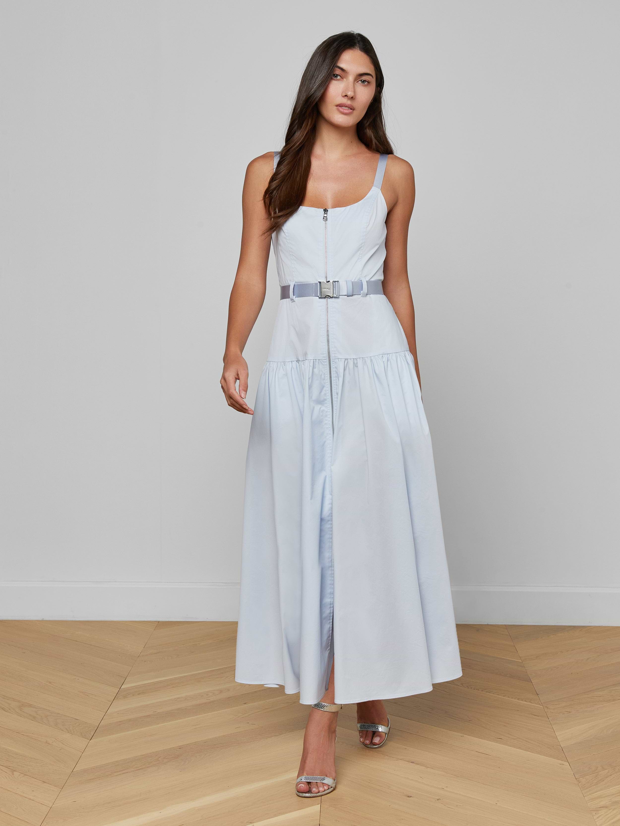 Wearing the Rima Maxi Dress—a pale blue, sleeveless, belted stretch poplin maxi dress—a woman paired with silver heels stands against a plain white background.