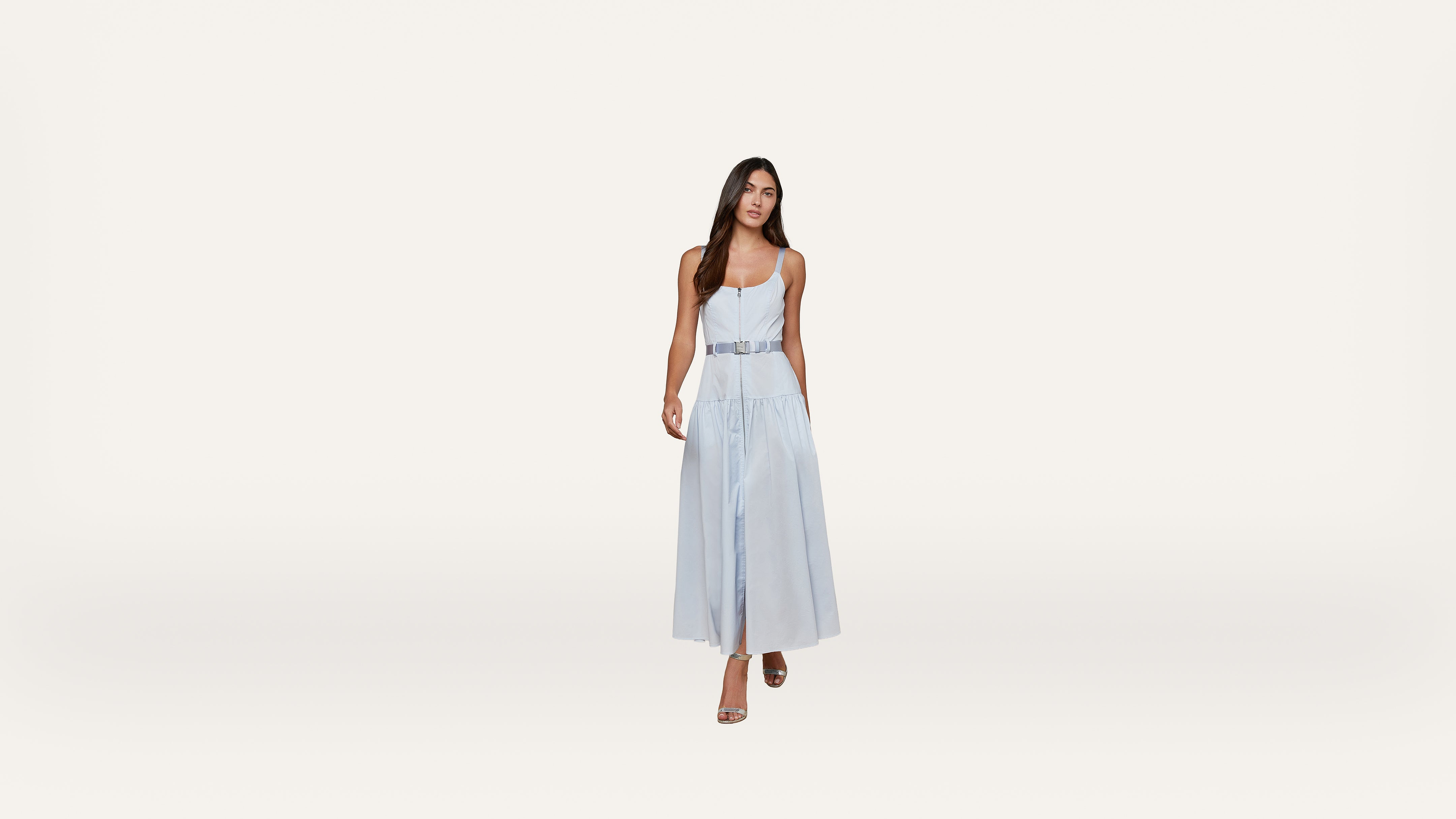 A woman wearing a light blue sleeveless dress and heels walks confidently against a plain white background.