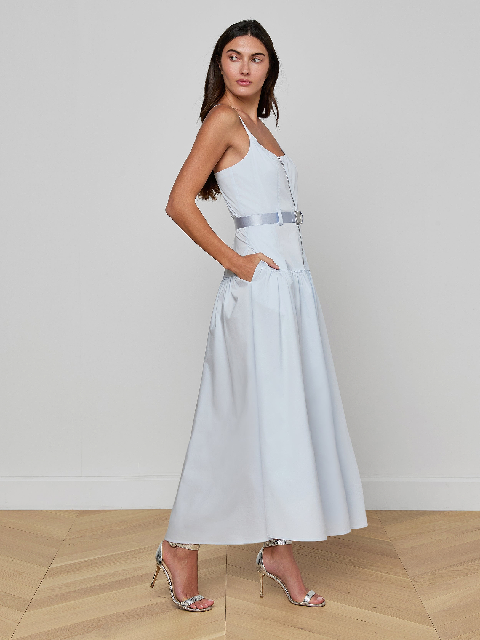 A woman stands sideways against a plain white background wearing the Rima Maxi Dress, a pale blue stretch poplin maxi style featuring a belt and pockets, paired with silver high-heeled sandals.