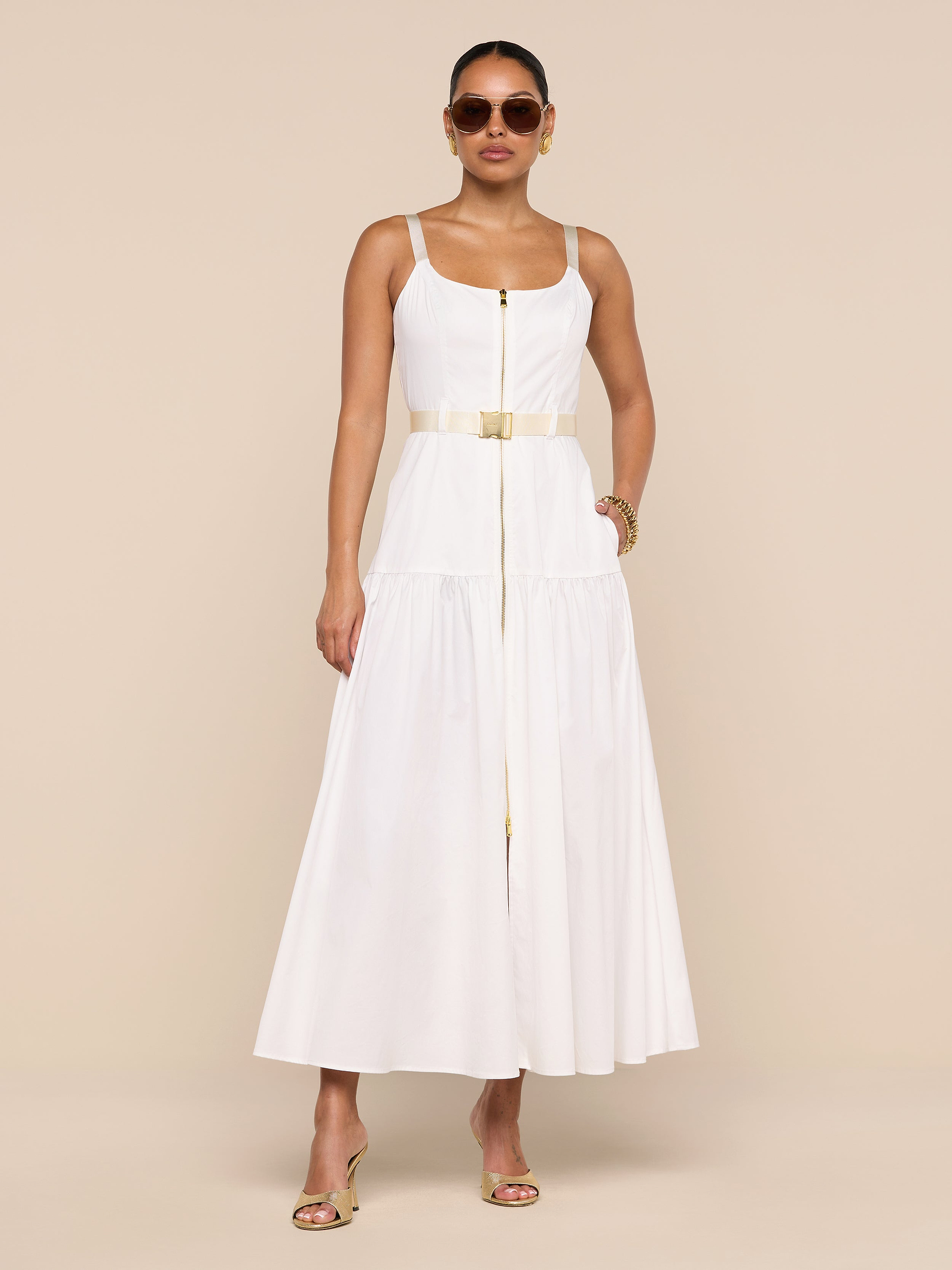 Wearing sunglasses, a woman stands against a plain white background with one hand in her pocket and the other relaxed by her side, modeling the Rima Dress—a sleeveless stretch poplin white maxi dress featuring an adjustable belt.