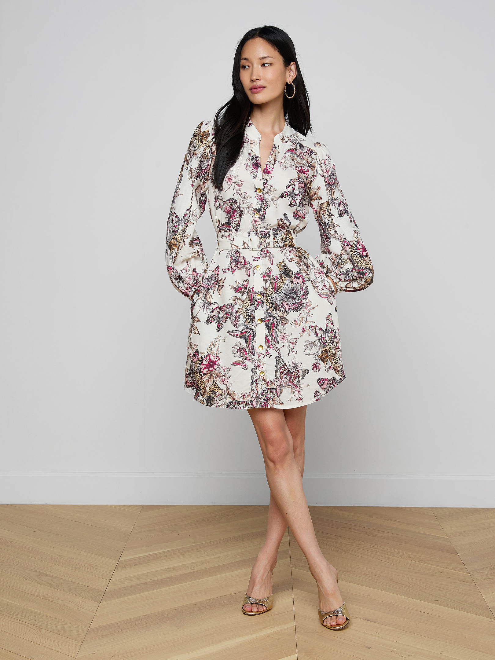 A woman stands against a plain background, wearing the Kiersten Dress—a vibrant print piece featuring long sleeves and a short hemline. She has long hair, hoop earrings, and is styled with open-toed heels.