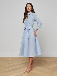 A woman stands against a plain white background wearing the Ralphie Midi Dress—a light blue poplin striped shirt dress with a waist tie—paired with nude heeled sandals.