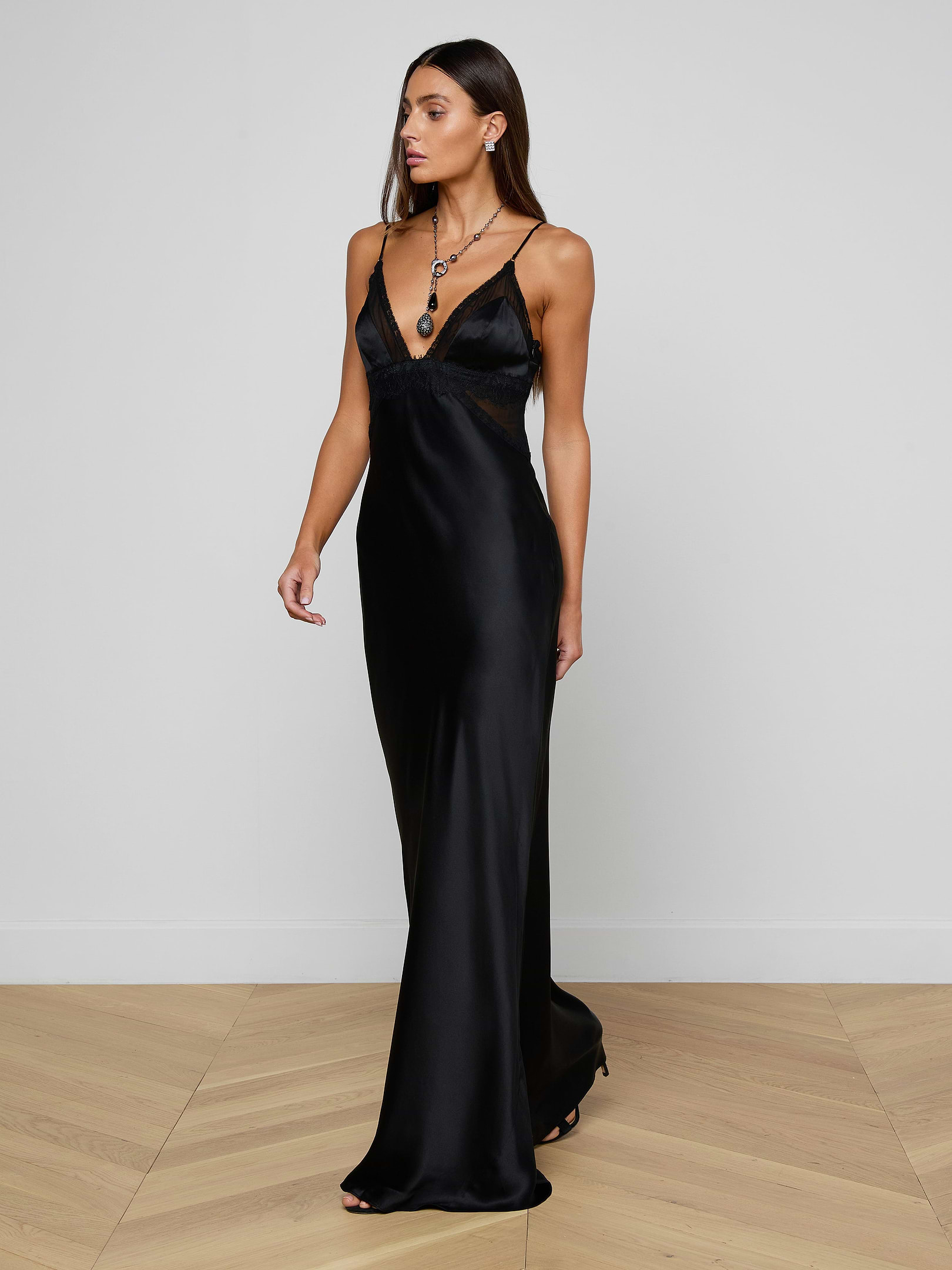 Wearing the Elsa Silk Gown—a long, sleeveless black slip dress with a V-neckline—a woman stands against a plain light background, looking to the left and accessorized with layered necklaces.