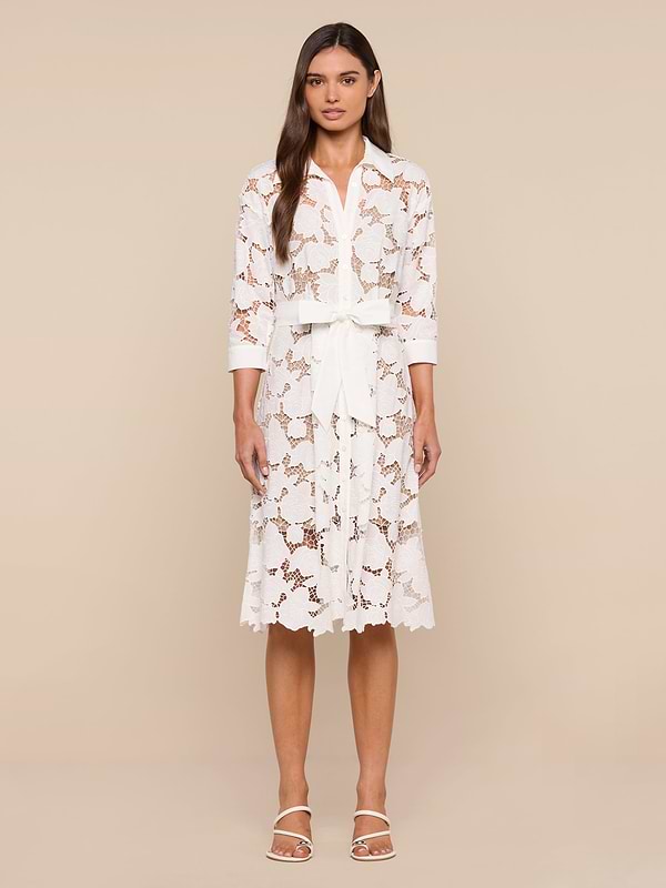A woman stands against a plain white background, wearing the May Eyelet Shirt Dress—a white cotton midi dress with three-quarter sleeves and a tied waist belt—paired with open-toed white sandals. She has long, straight hair and a neutral expression.