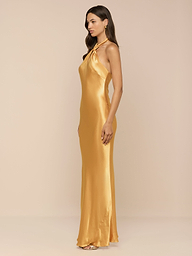 A woman stands in profile against a plain white background, wearing the Saphara Halter Gown—a long, gold satin dress.