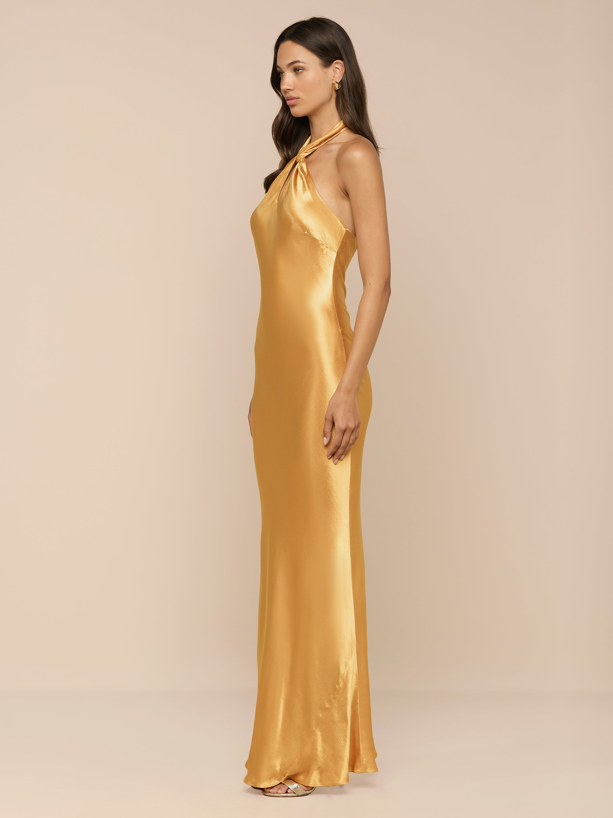 A woman stands in profile against a plain white background, wearing the Saphara Halter Gown—a long, gold satin dress.