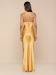 A woman with long brown hair wears the Saphara Halter Gown—a gold satin dress featuring a built-in bra—and stands facing away against a plain white background.