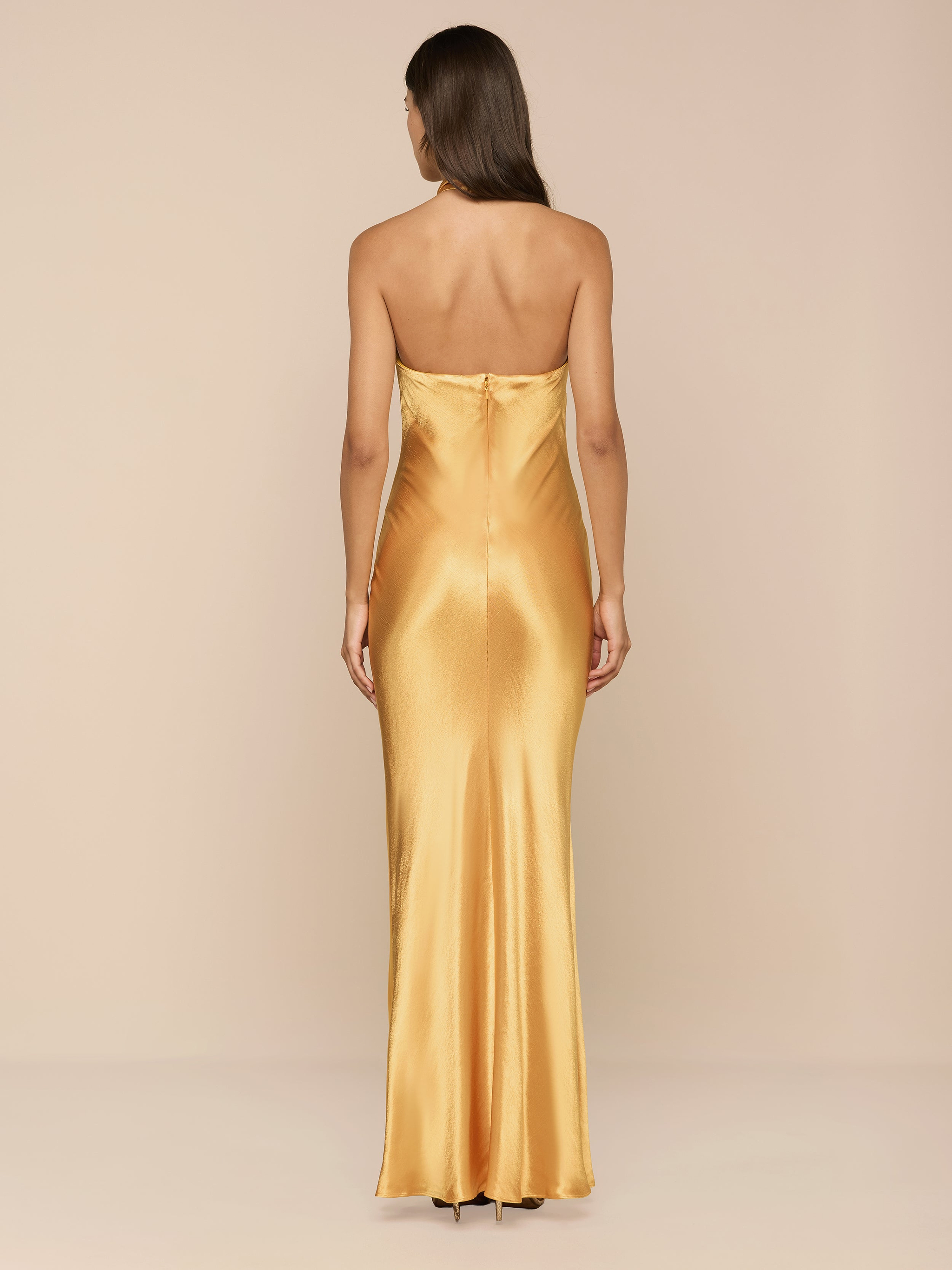 A woman with long brown hair wears the Saphara Halter Gown—a gold satin dress featuring a built-in bra—and stands facing away against a plain white background.