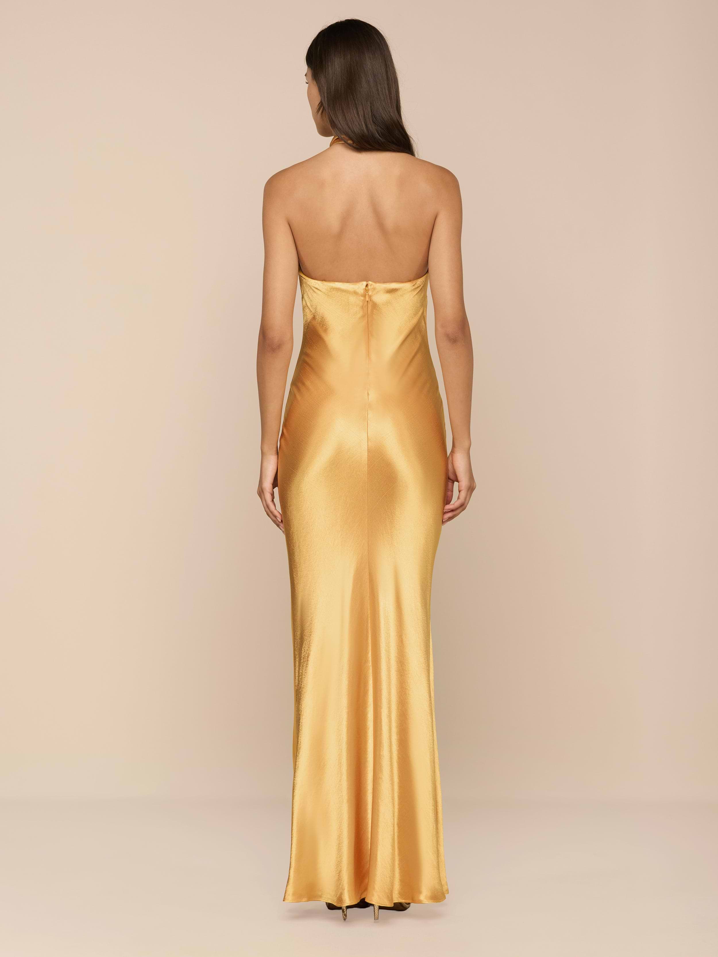 A woman with long brown hair wears the Saphara Halter Gown—a gold satin dress featuring a built-in bra—and stands facing away against a plain white background.
