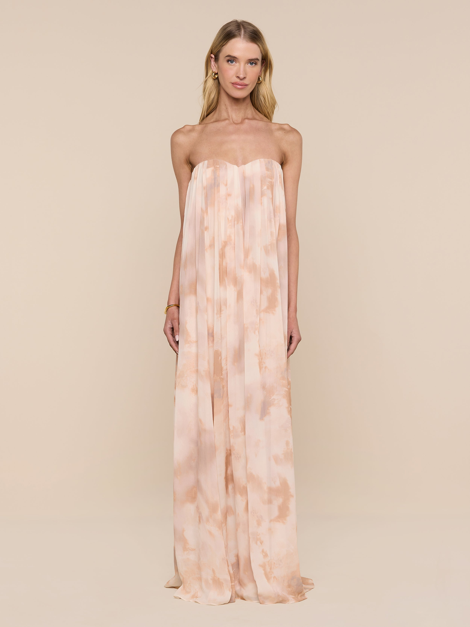 A woman stands against a plain white background, wearing the Raine Strapless Gown in a light peach and cream tie-dye design. The floor-length dress features a built-in corset, and she faces forward with a neutral expression, her arms at her sides.