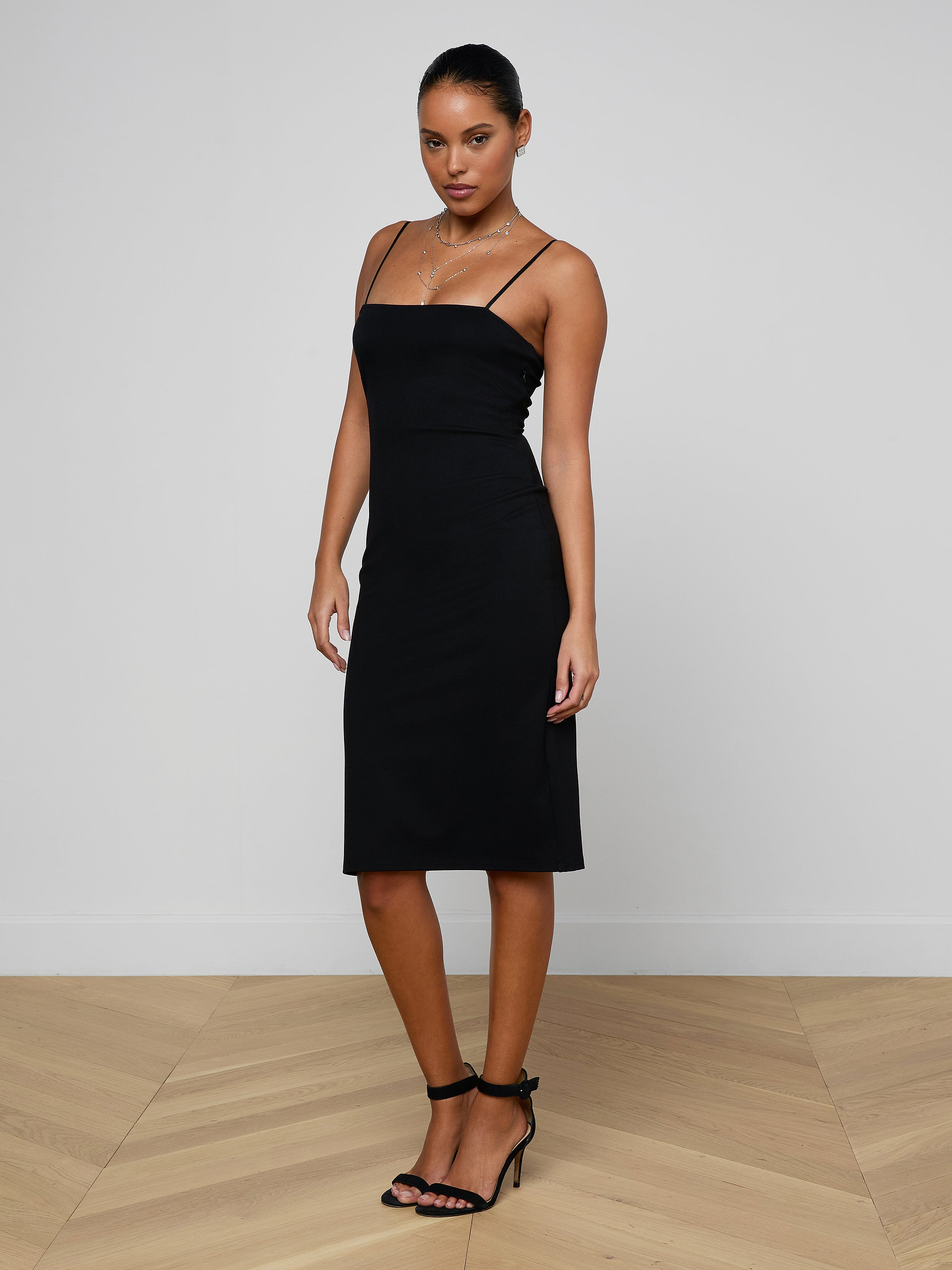 Wearing the Eira Midi Dress, a woman poses against a plain white background, looking at the camera with one hand by her side. The black knee-length slip dress features adjustable straps and is paired with black heels.