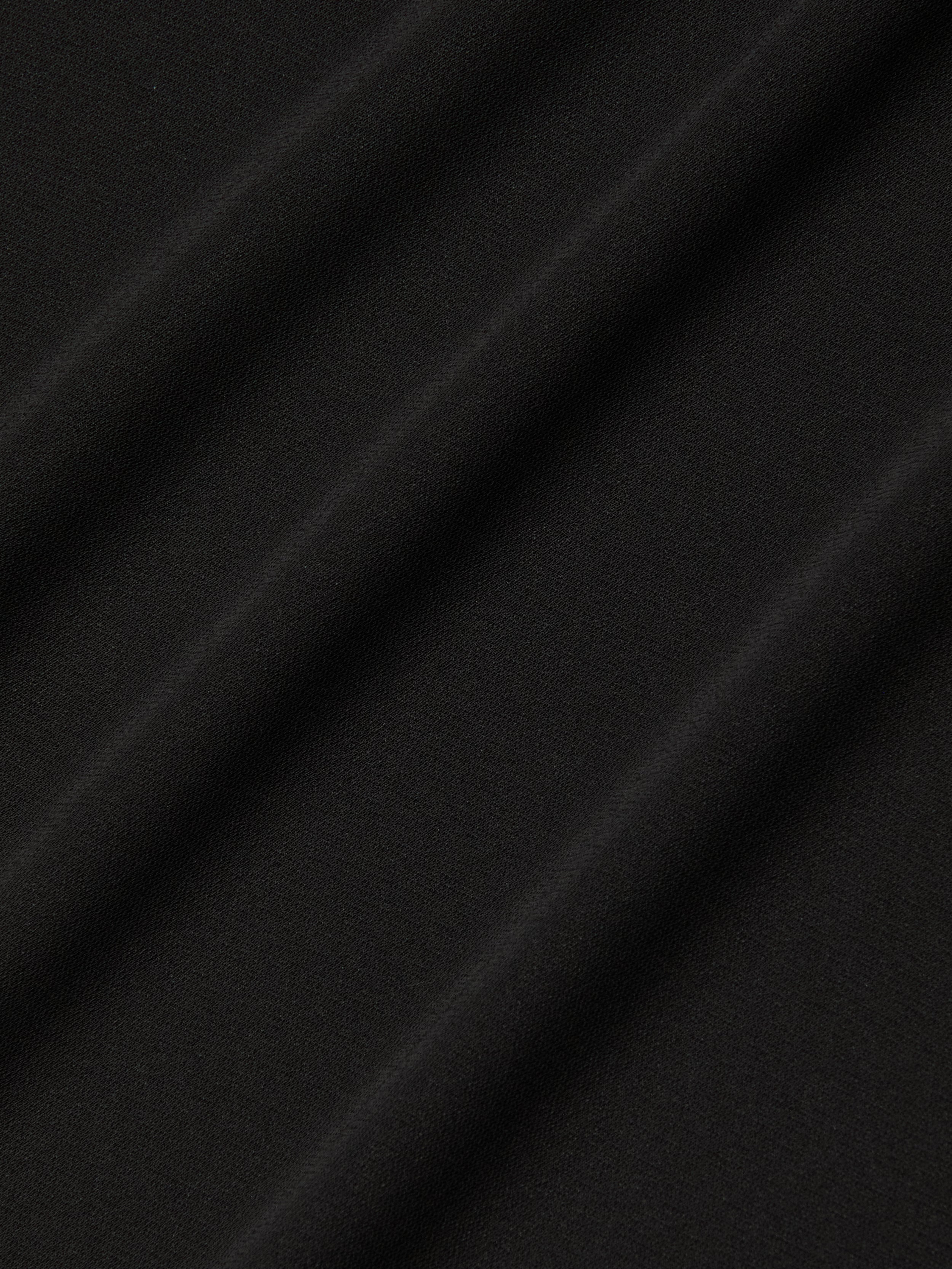 Close-up of the Raye Slip Lining Dress in black stretch jersey fabric, featuring diagonal, evenly spaced folds that create a soft and subtle textured pattern. The surface appears smooth and matte, with shadows accentuating the gentle contours of the dress’s pleats.