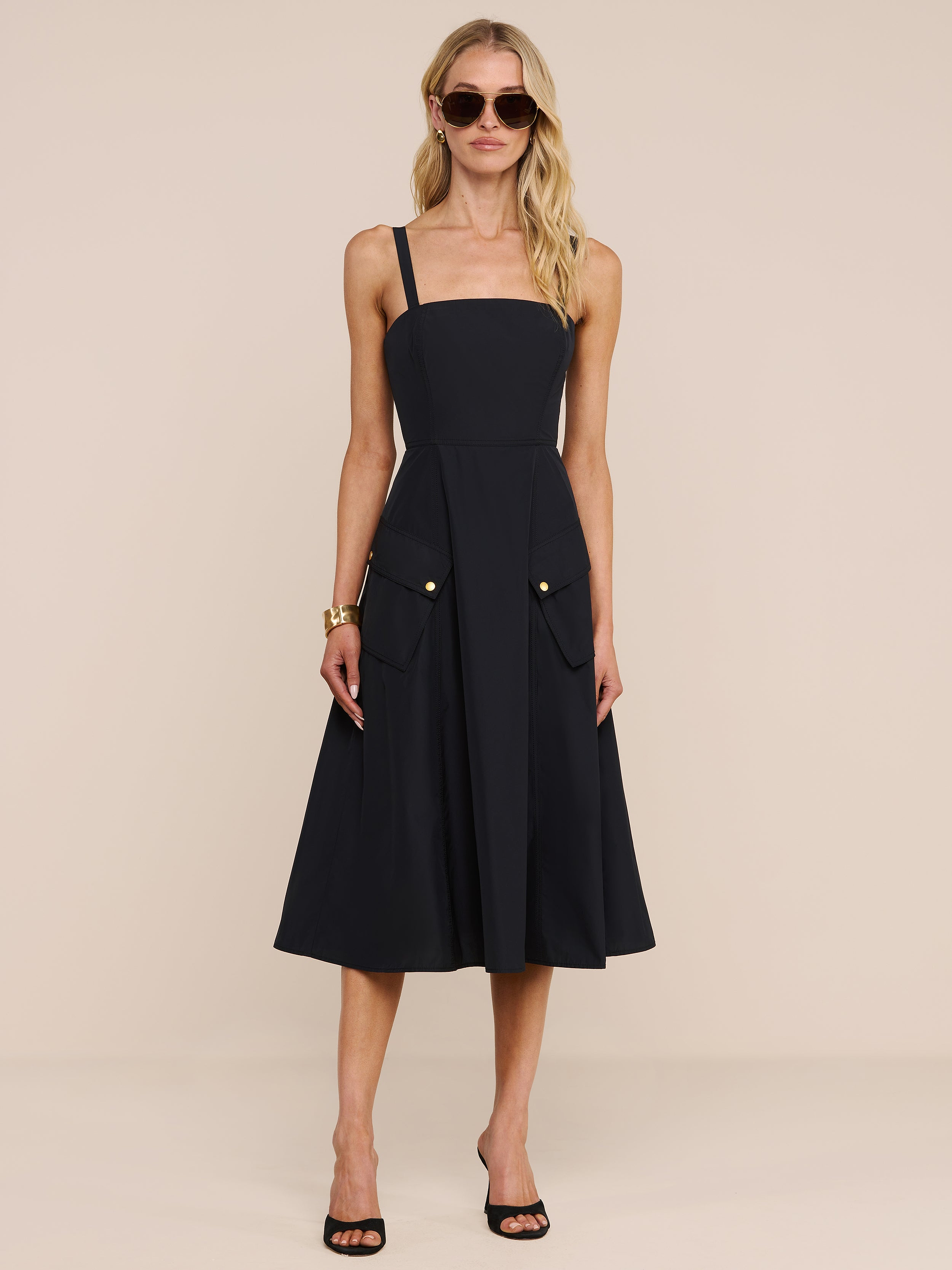 A woman wearing the Ayela Midi Dress, featuring a black midi length and square neckline with front pockets adorned with gold buttons, pairs it with black heels and sunglasses while standing against a plain white background.
