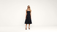 A woman wearing sunglasses and a sleeveless black midi dress with spaghetti straps and black heels stands against a plain white background.