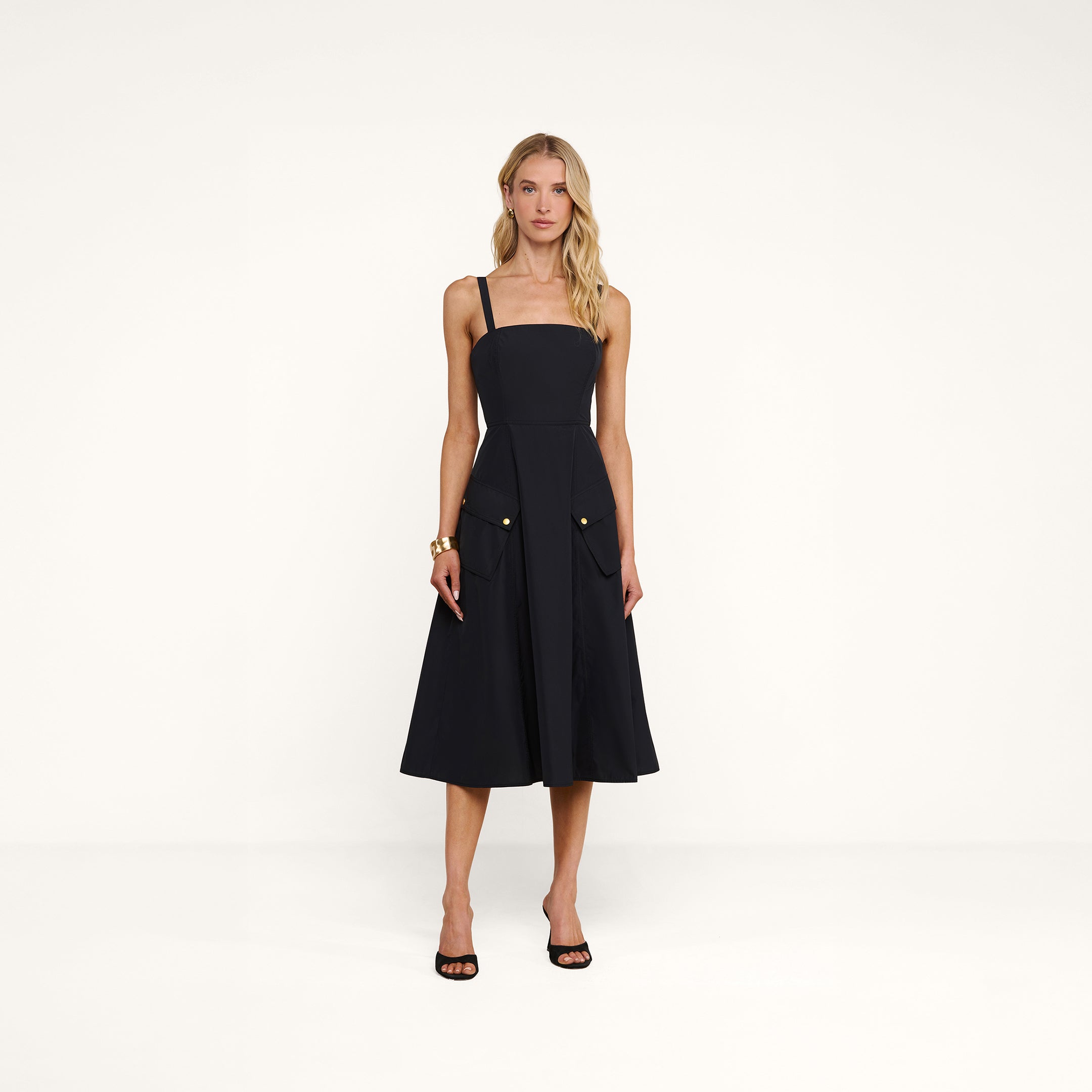 A woman stands in a studio wearing the Ayela Midi Dress—a sleeveless, knee-length black dress with a square neckline, wide straps, and large front pockets. She pairs it with black heeled sandals, her long wavy blonde hair framing her neutral expression.