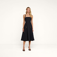 A woman stands in a studio wearing the Ayela Midi Dress—a sleeveless, knee-length black dress with a square neckline, wide straps, and large front pockets. She pairs it with black heeled sandals, her long wavy blonde hair framing her neutral expression.