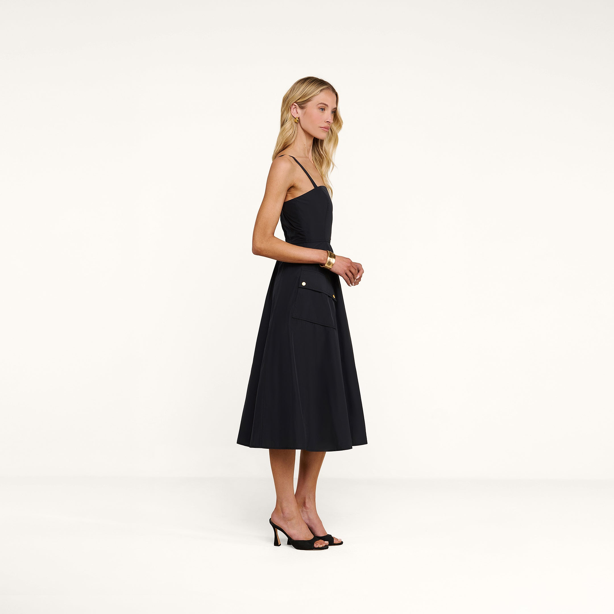 A woman stands in profile against a plain white background, wearing the Ayela Midi Dress—a black sleeveless midi dress with a square neckline—paired with black heels.