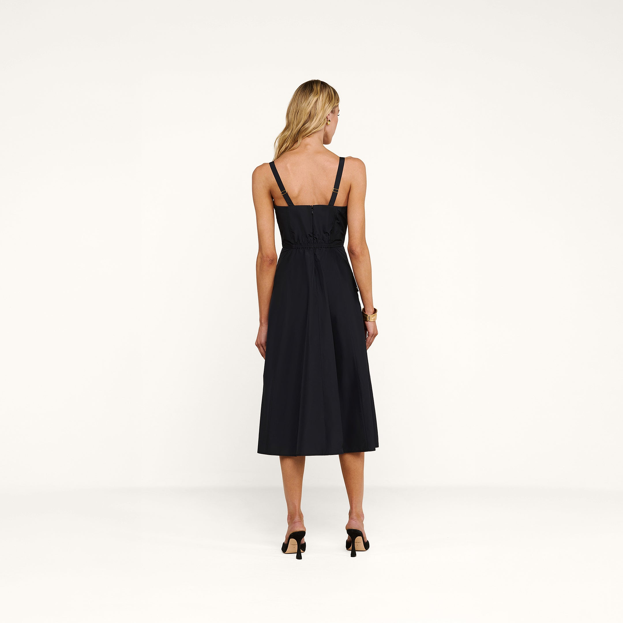 A woman stands with her back to the camera, wearing the Ayela Midi Dress—a sleeveless black midi dress with removable straps—paired with black heels, against a plain white background.