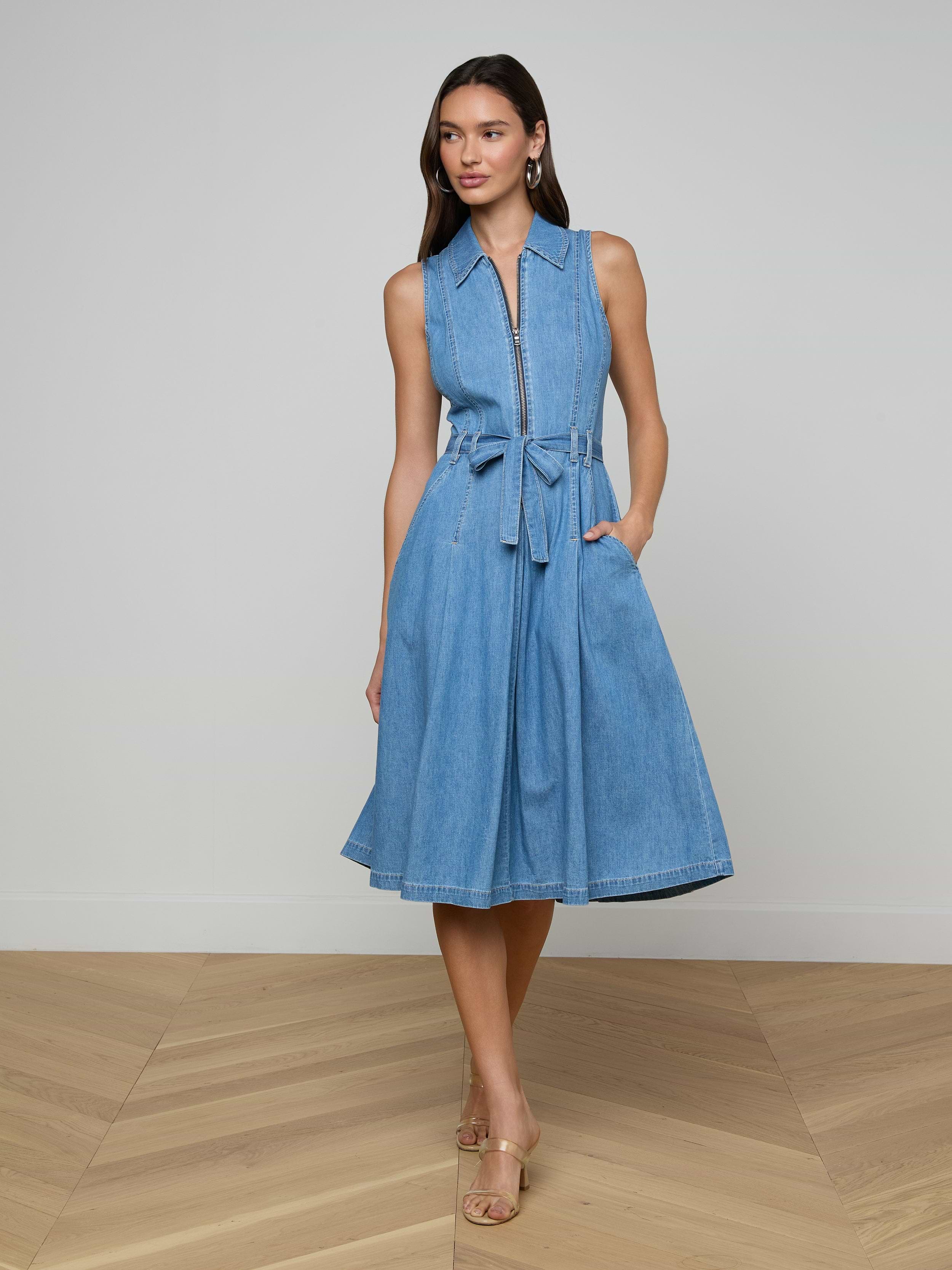 A woman stands wearing the Kieran Shirt Dress—a sleeveless, knee-length, light blue featherweight denim dress with a tie belt and pockets—paired with tan heels. She poses with her hands in her pockets against a plain, light background.