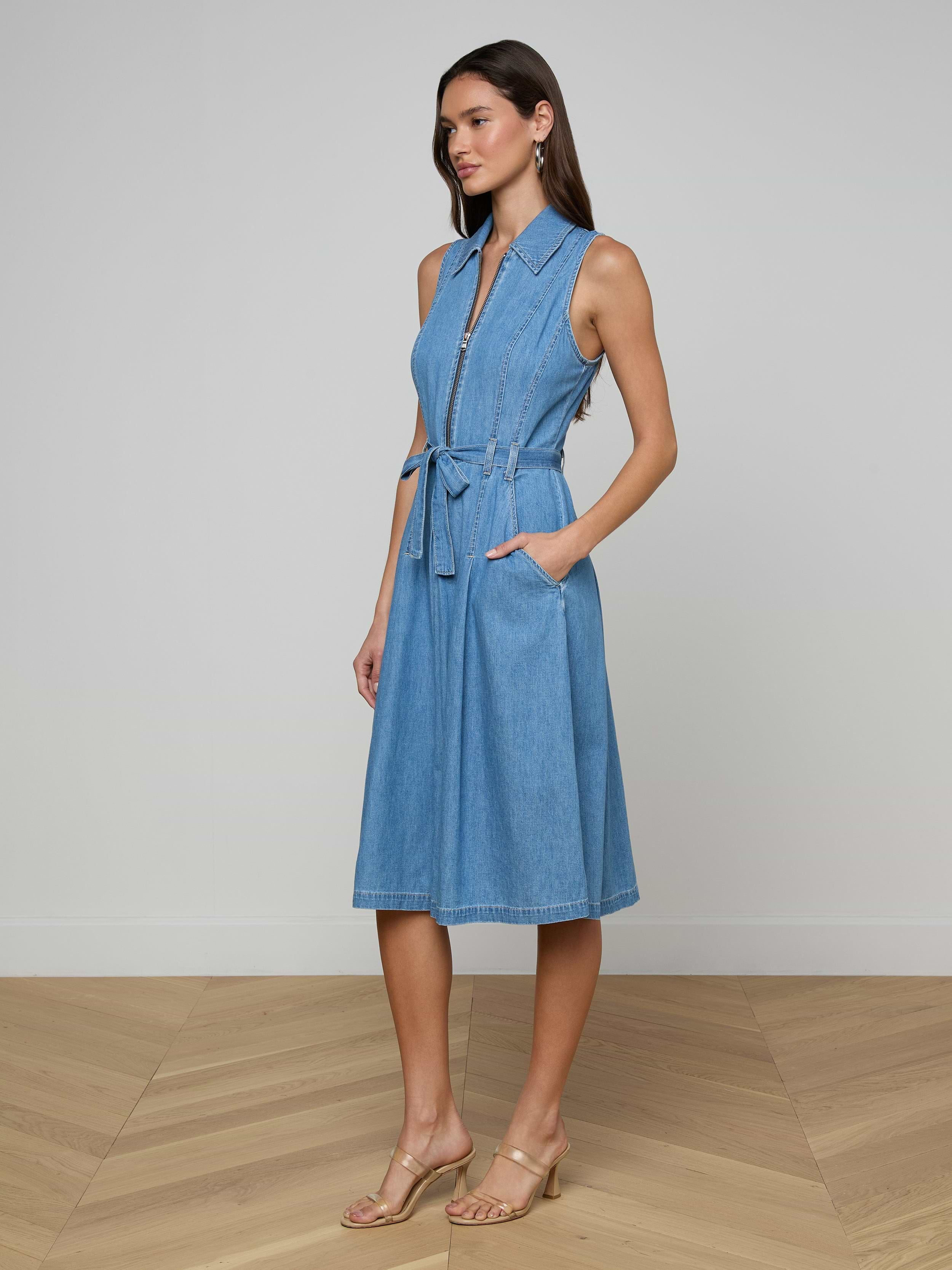 A woman stands wearing the Kieran Shirt Dress, a sleeveless, knee-length featherweight denim dress featuring a tie belt and pockets. She has long hair and is styled in heeled sandals against a plain white background.