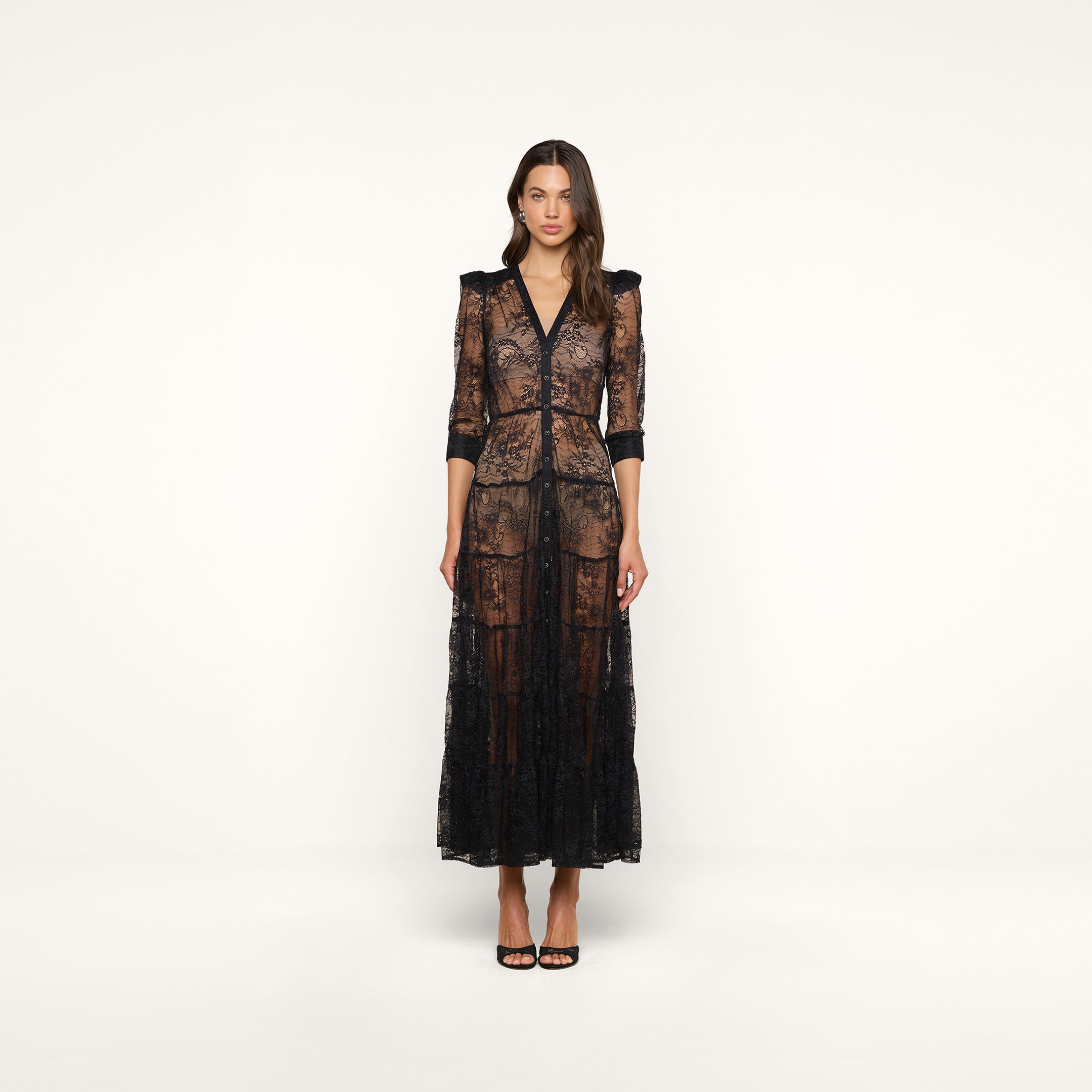 A woman stands against a plain background wearing the Seraphina Lace Tiered Maxi Dress, featuring sheer lace fabric, 3/4 length sleeves, and styled with black high-heeled sandals.