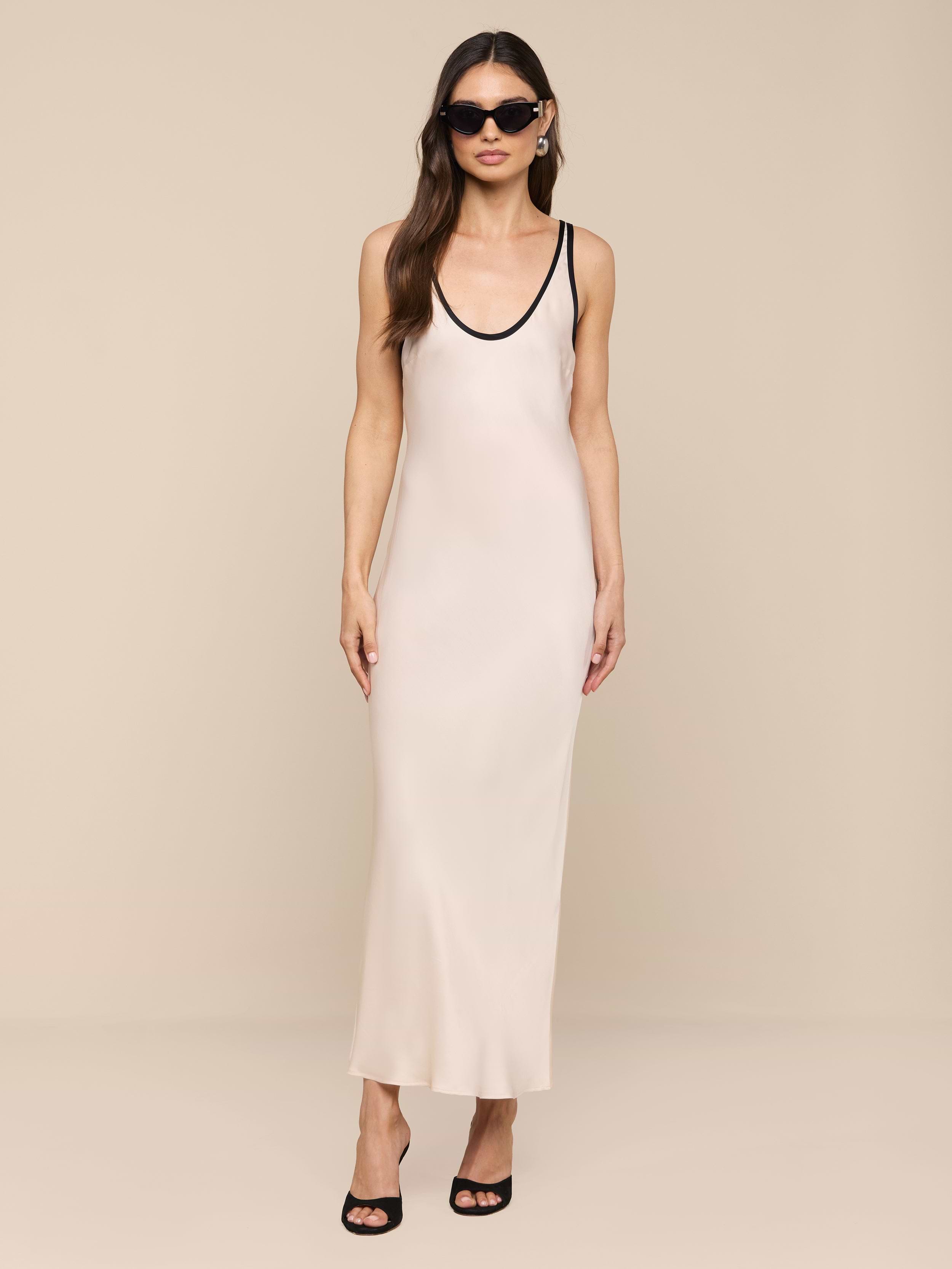 A woman with long hair stands against a plain white background wearing the Akiya Satin Maxi Dress, a long, vintage-inspired white bias-cut satin dress. She accessorizes with black sunglasses and black high-heeled shoes.