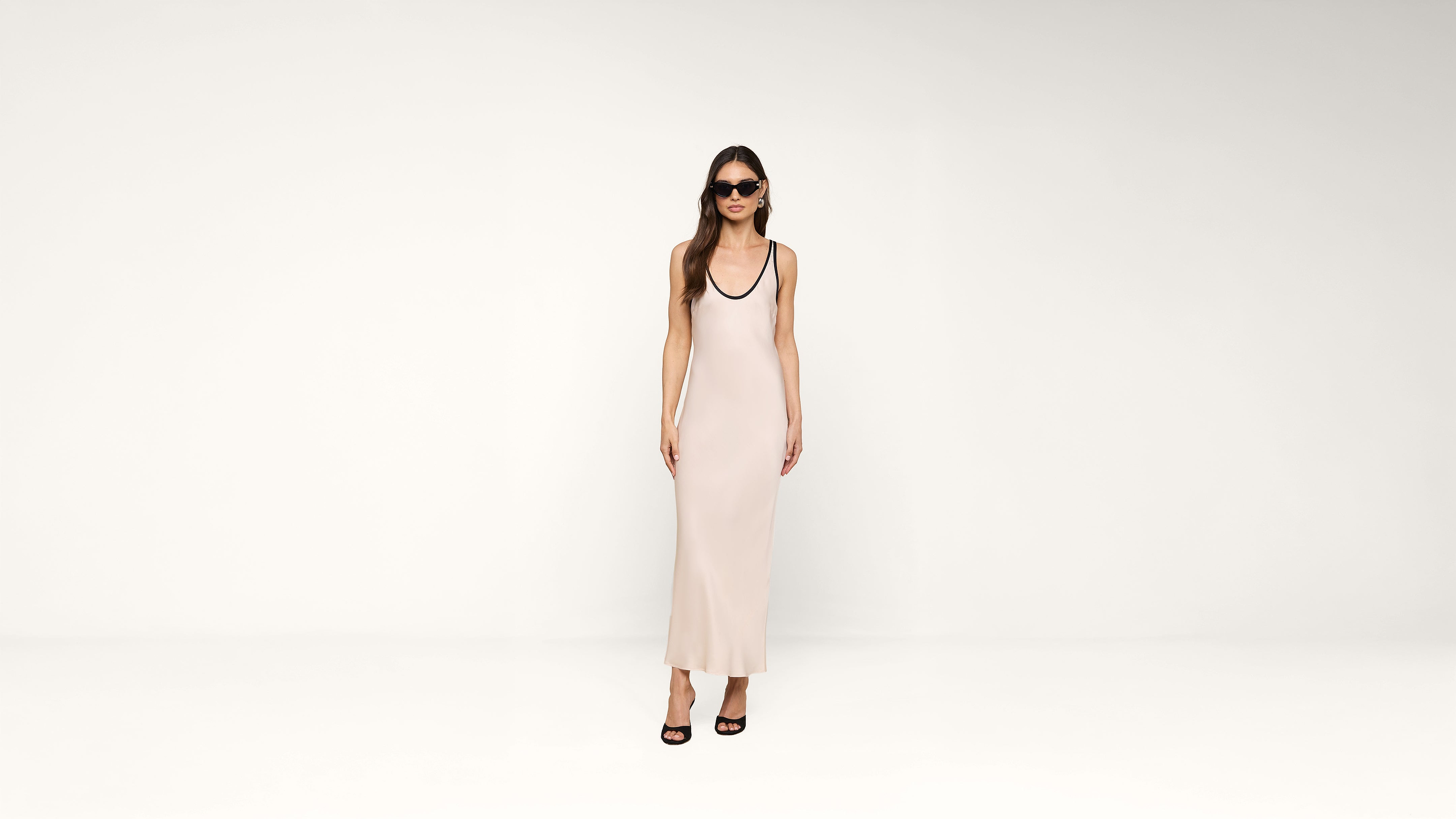 A woman in sunglasses and a long, pale pink sleeveless dress stands against a plain white background, wearing black high-heeled sandals.