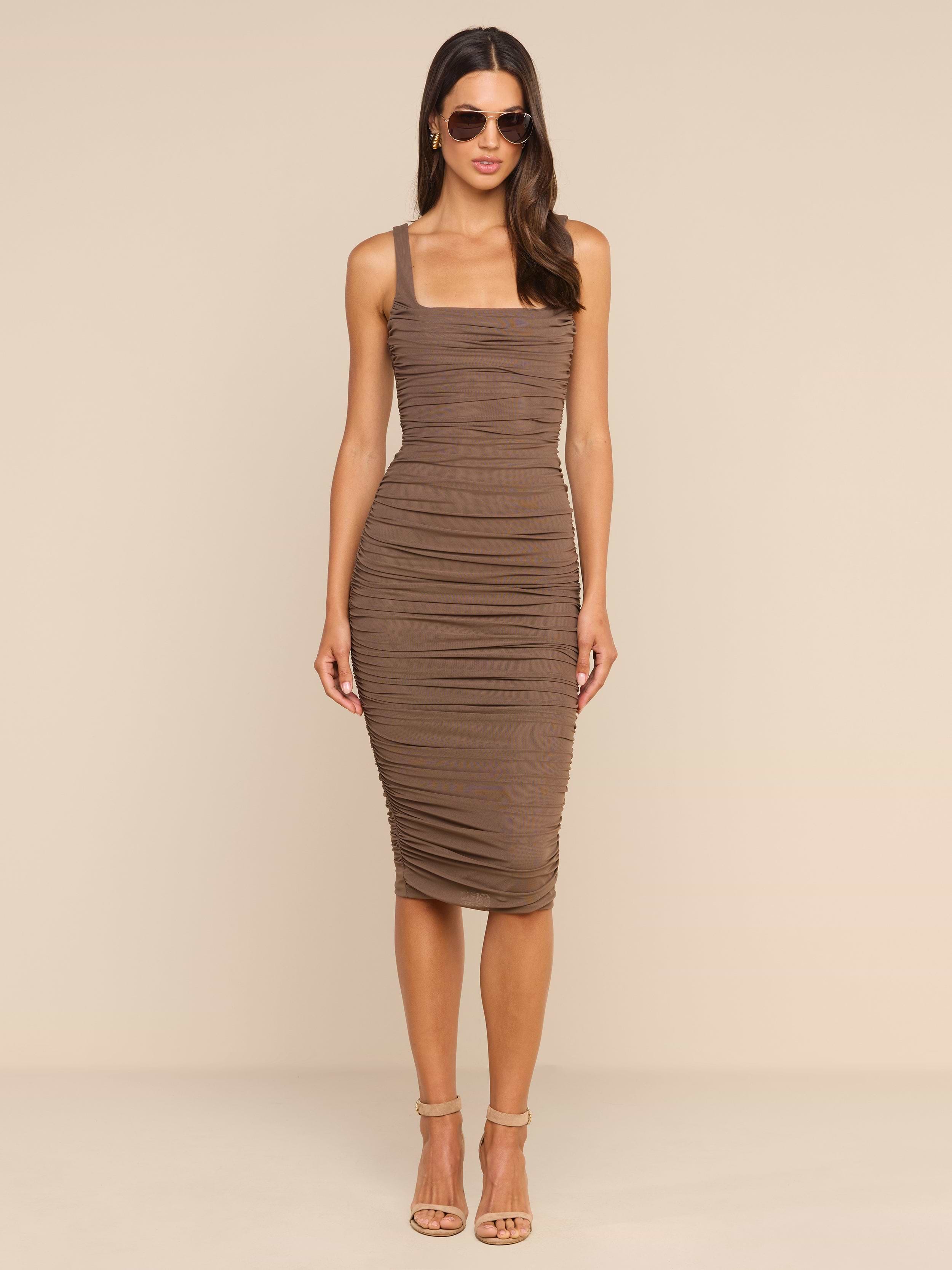 A woman with long hair, wearing sunglasses and open-toed heels, stands facing forward against a plain white background. She is dressed in the Anya Shirred Dress, a form-hugging brown midi dress with ruching details.