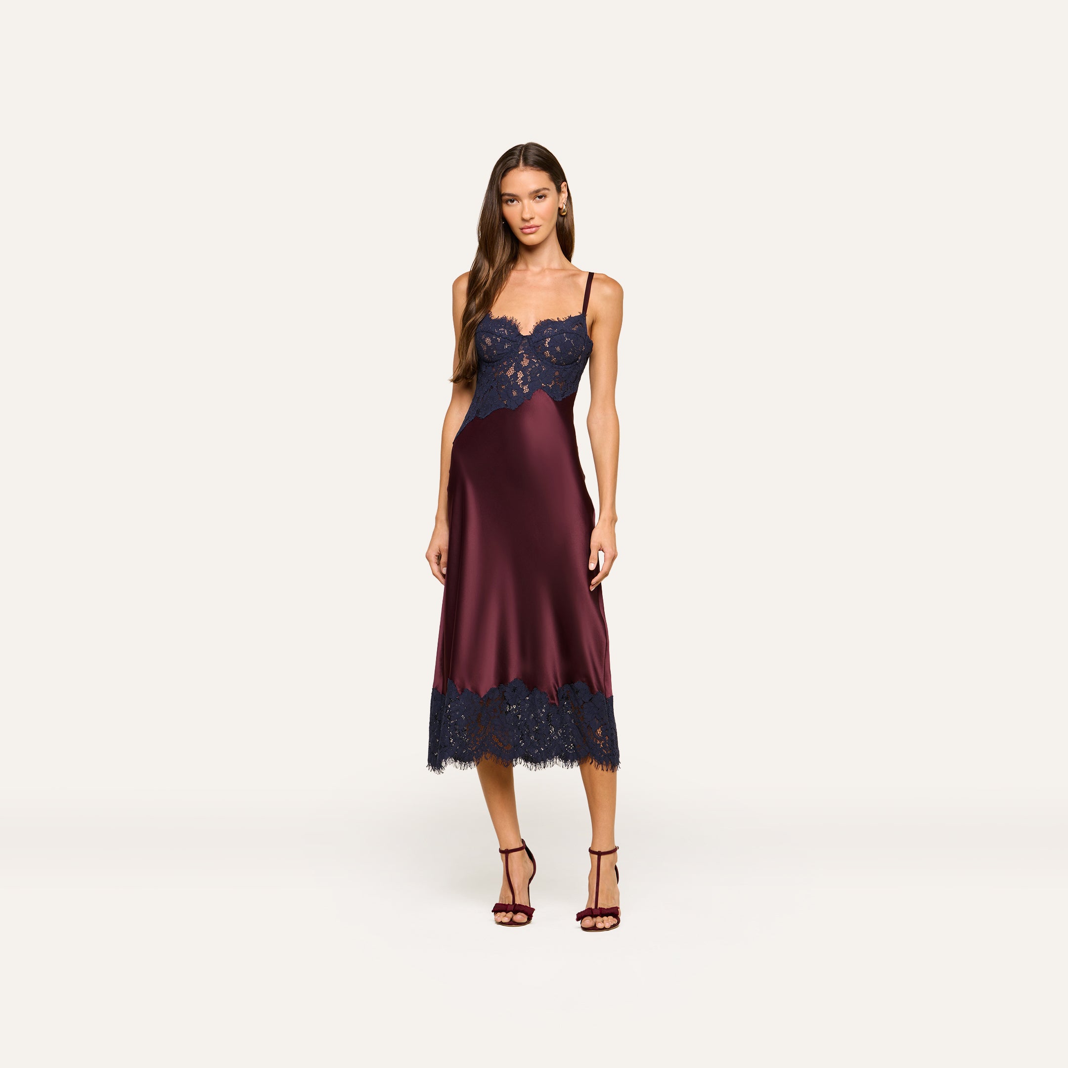 A woman wearing the Araminta Lace Dress—a burgundy silk midi dress with navy lace appliqués and spaghetti straps—paired with matching burgundy heels, stands against a plain light background.