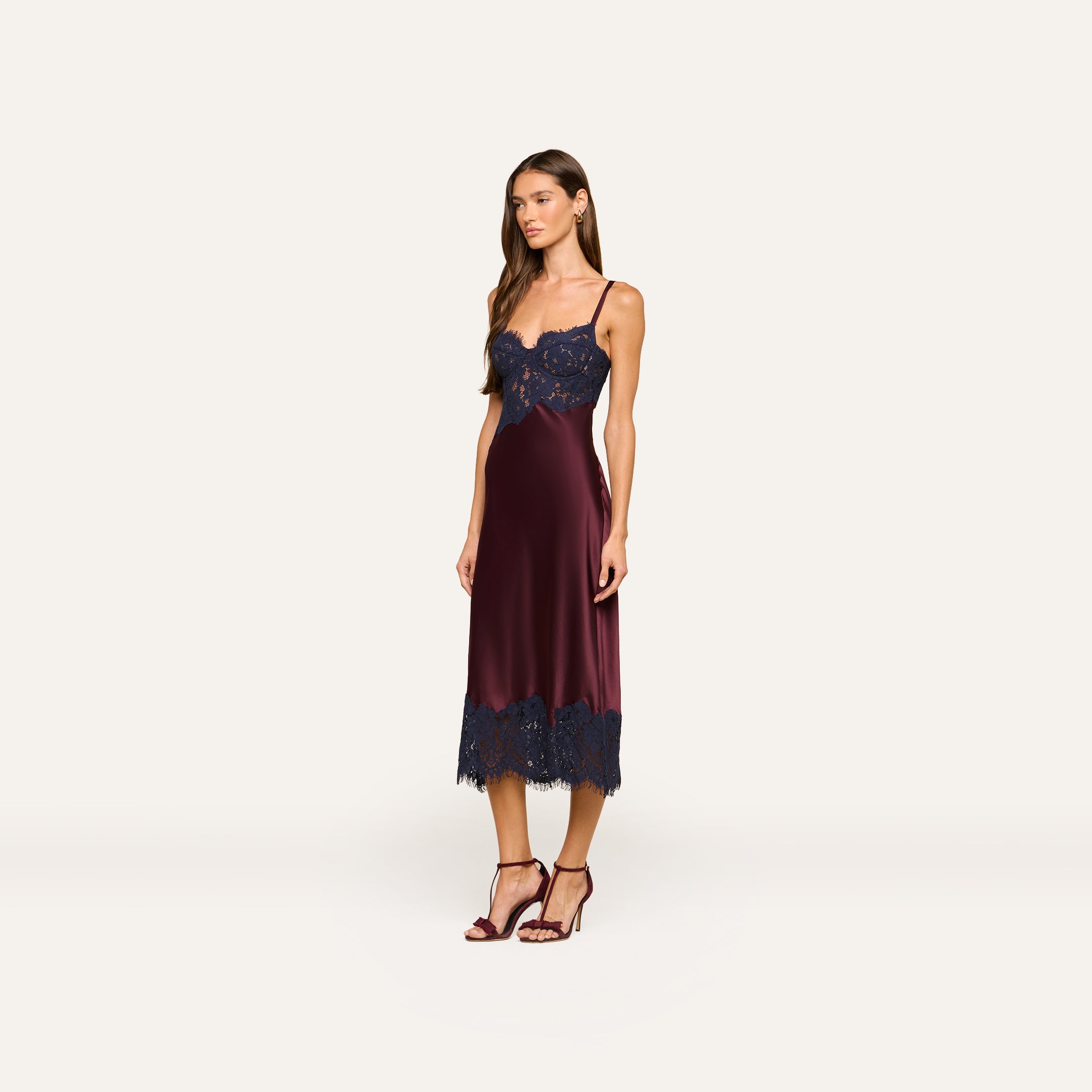 A woman wearing the Araminta Lace Dress—featuring burgundy silk with navy lace appliqués on the bodice and hem—stands in heels against a plain light background.