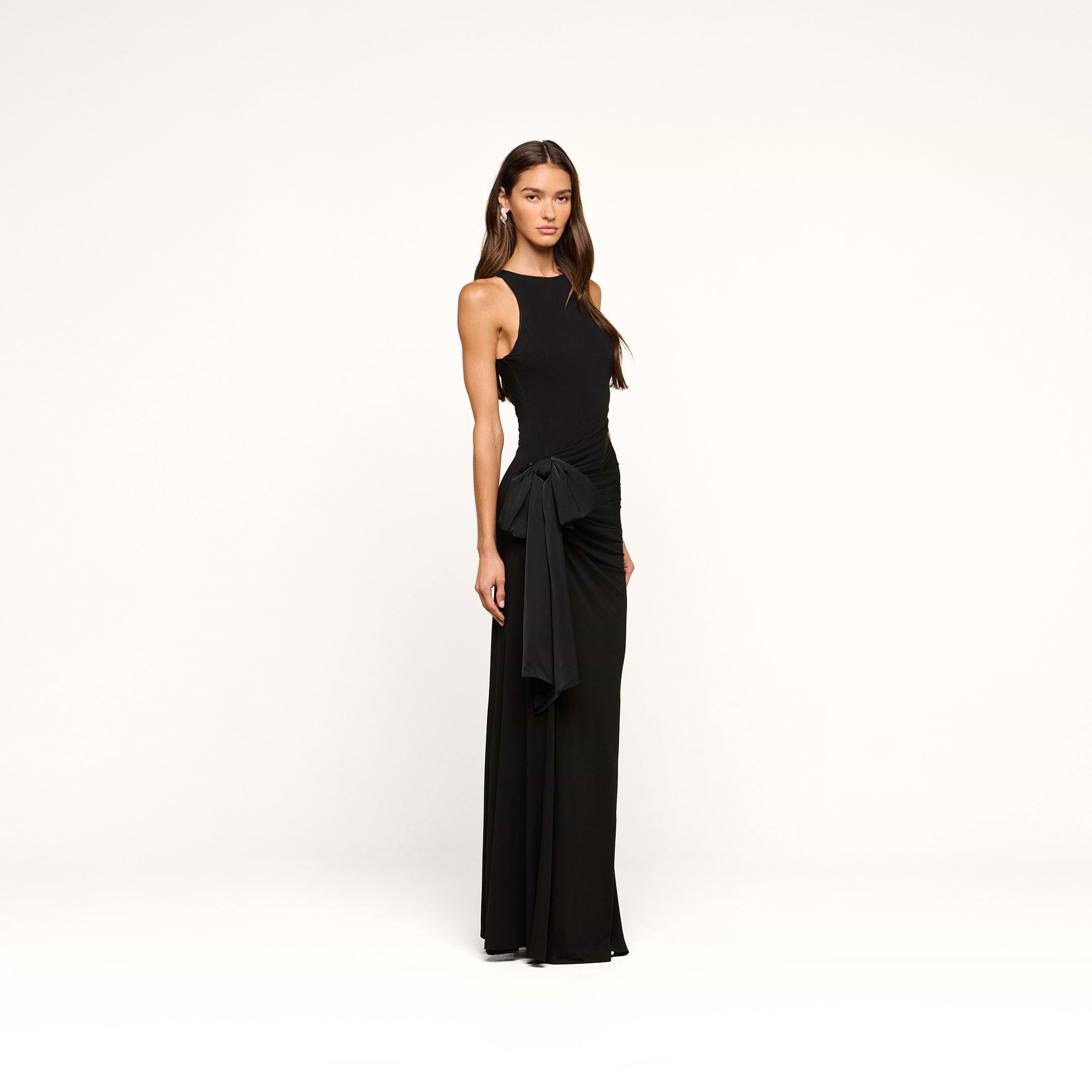 A woman stands in the Frost Gown, a sleeveless, floor-length black tie dress featuring a side sash detail, posing against a plain white background.