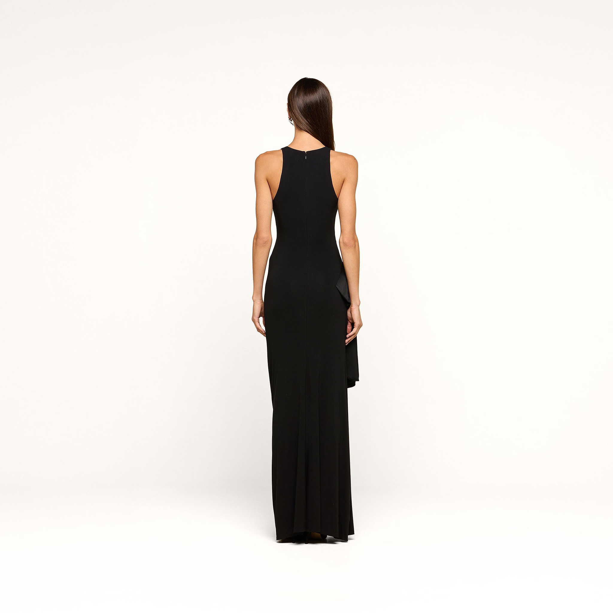 A woman with long brown hair wears the Frost Gown—a sleeveless, floor-length black tie dress—standing in a minimal white studio, exuding an elegance worthy of New York Fashion Week.