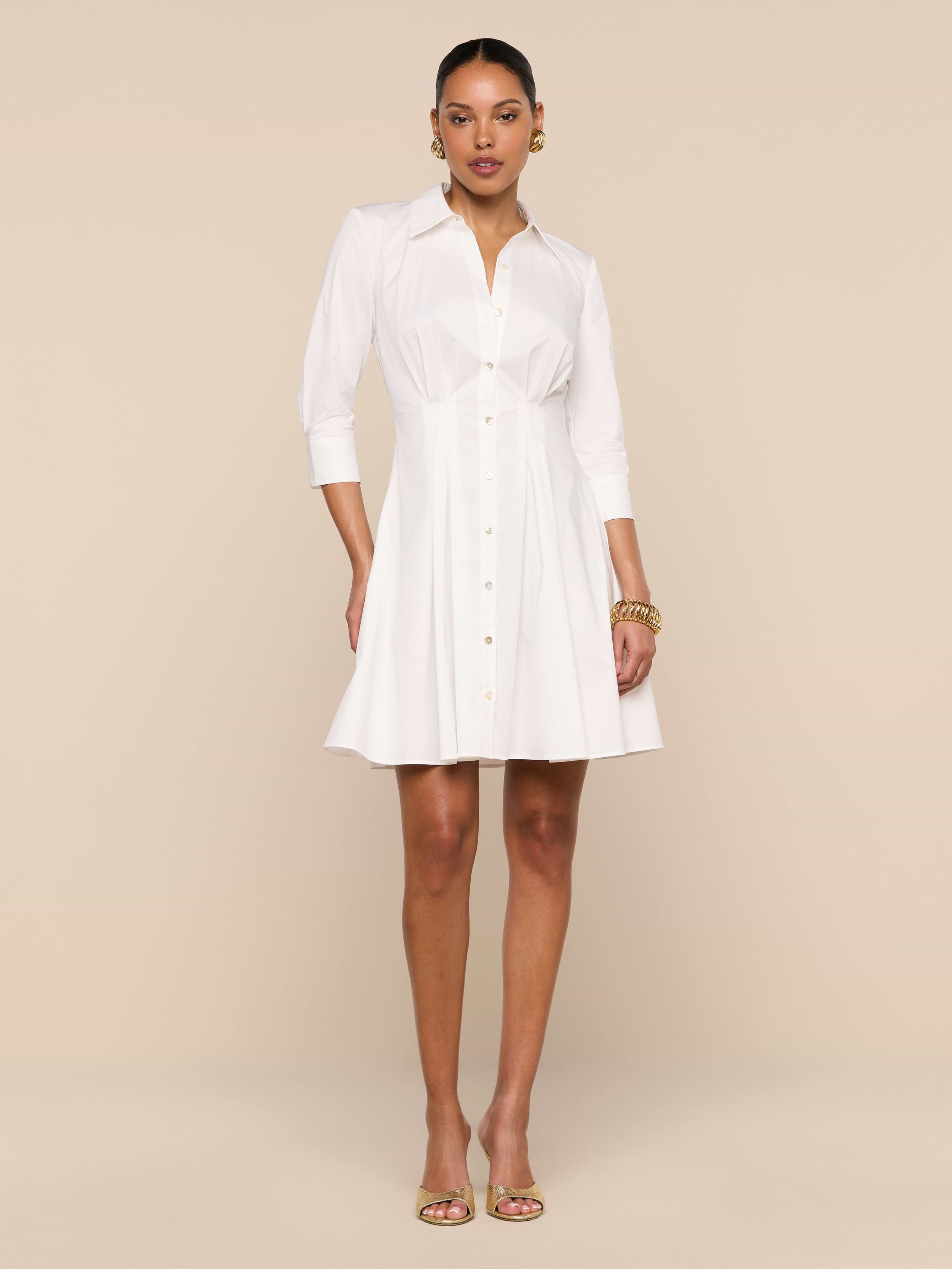 A woman stands against a plain white background wearing the Journi Shirt Dress—a white, button-down, knee-length design with three-quarter sleeves. She accessorizes with gold jewelry and open-toe heels, her left hand in her pocket to highlight the timeless silhouette of this cotton-blend poplin piece.