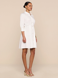 A woman stands in profile against a plain white background, wearing the Journi Shirt Dress—a light-colored cotton-blend poplin design with three-quarter sleeves—paired with gold heels. The timeless silhouette of the dress creates an effortlessly elegant look.