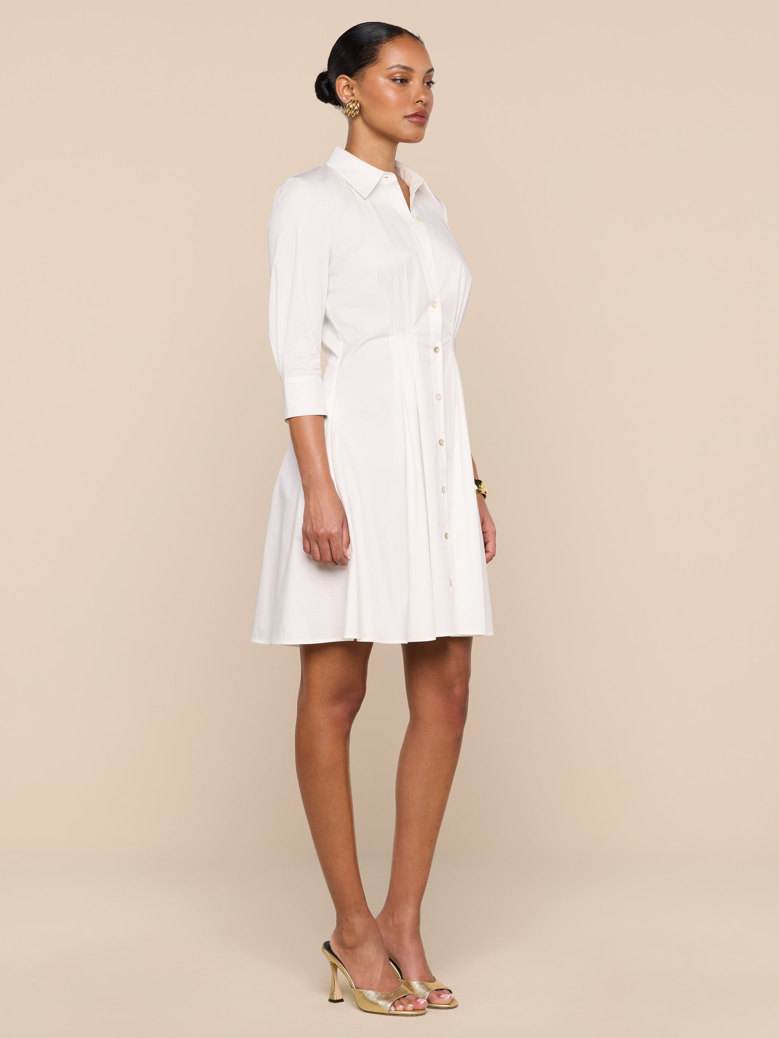 A woman stands in profile against a plain white background, wearing the Journi Shirt Dress—a light-colored cotton-blend poplin design with three-quarter sleeves—paired with gold heels. The timeless silhouette of the dress creates an effortlessly elegant look.