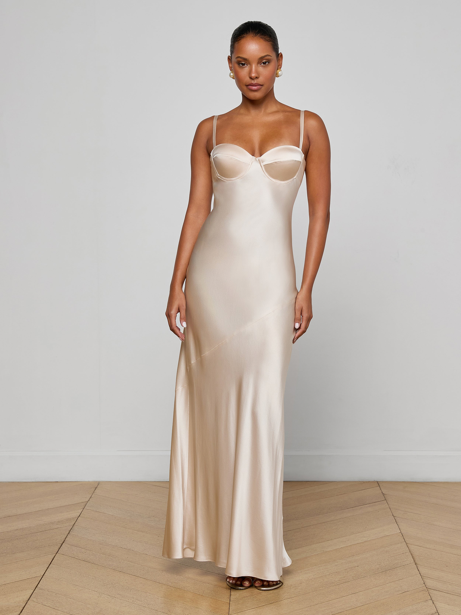A woman stands against a plain white background, wearing the Sayla Silk Gown—a long, sleeveless champagne dress with thin straps and a fitted bodice that evokes vintage glamour.