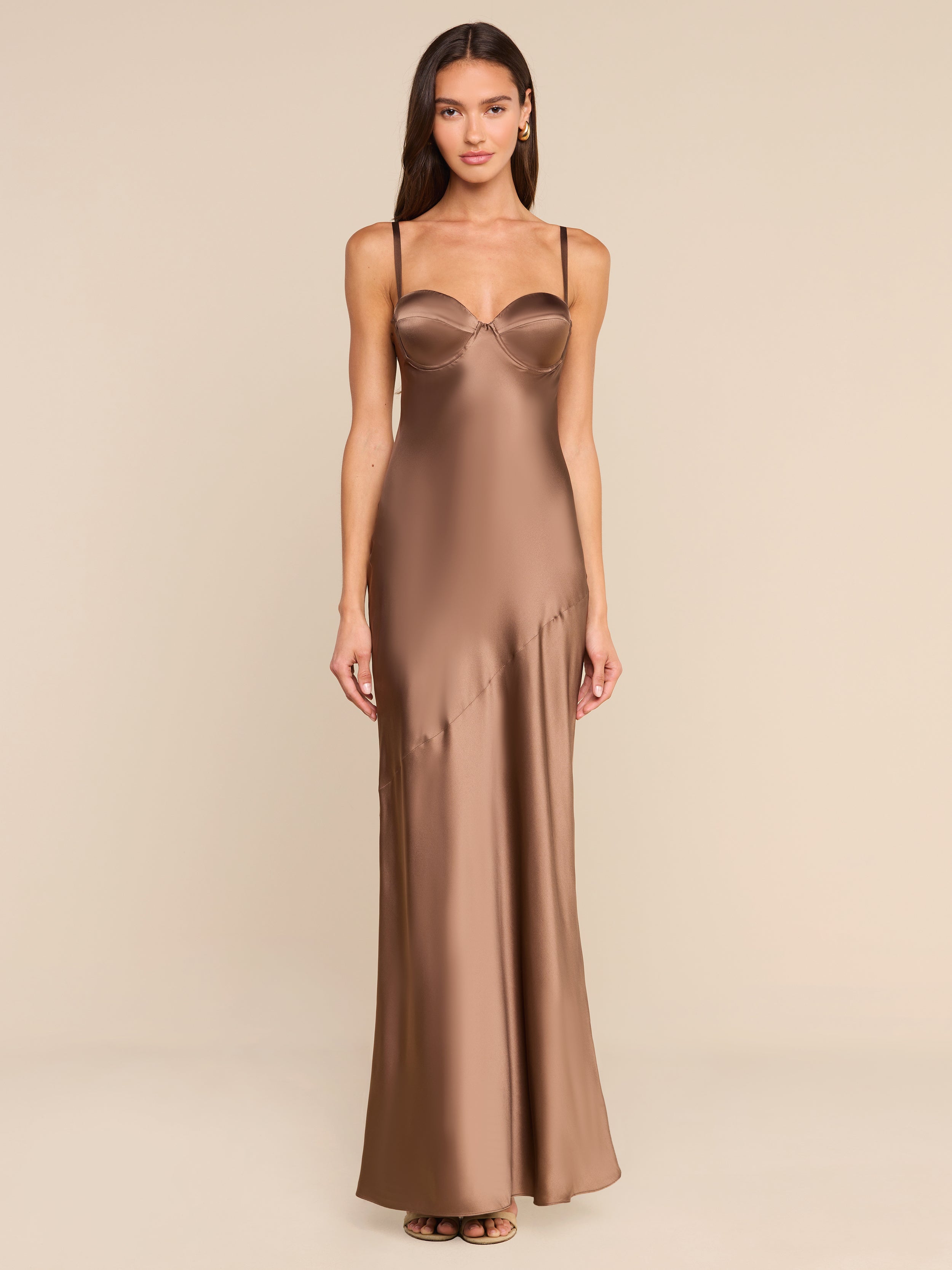 A woman stands facing forward in a plain, white studio setting, wearing the Sayla Silk Gown—a long bronze dress with thin straps.
