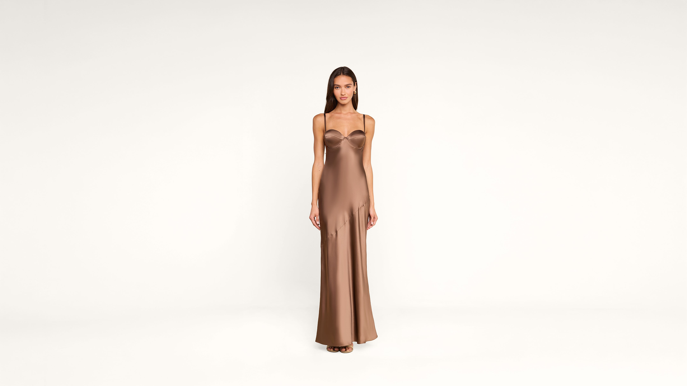 A woman stands facing forward, wearing a long, form-fitting, brown satin dress with thin straps against a plain white background.