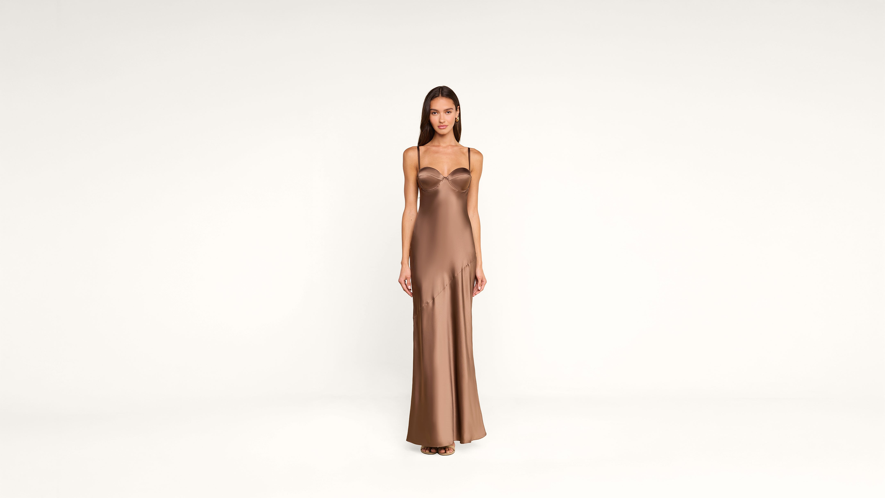 A woman stands facing forward, wearing a long, form-fitting, brown satin dress with thin straps against a plain white background.