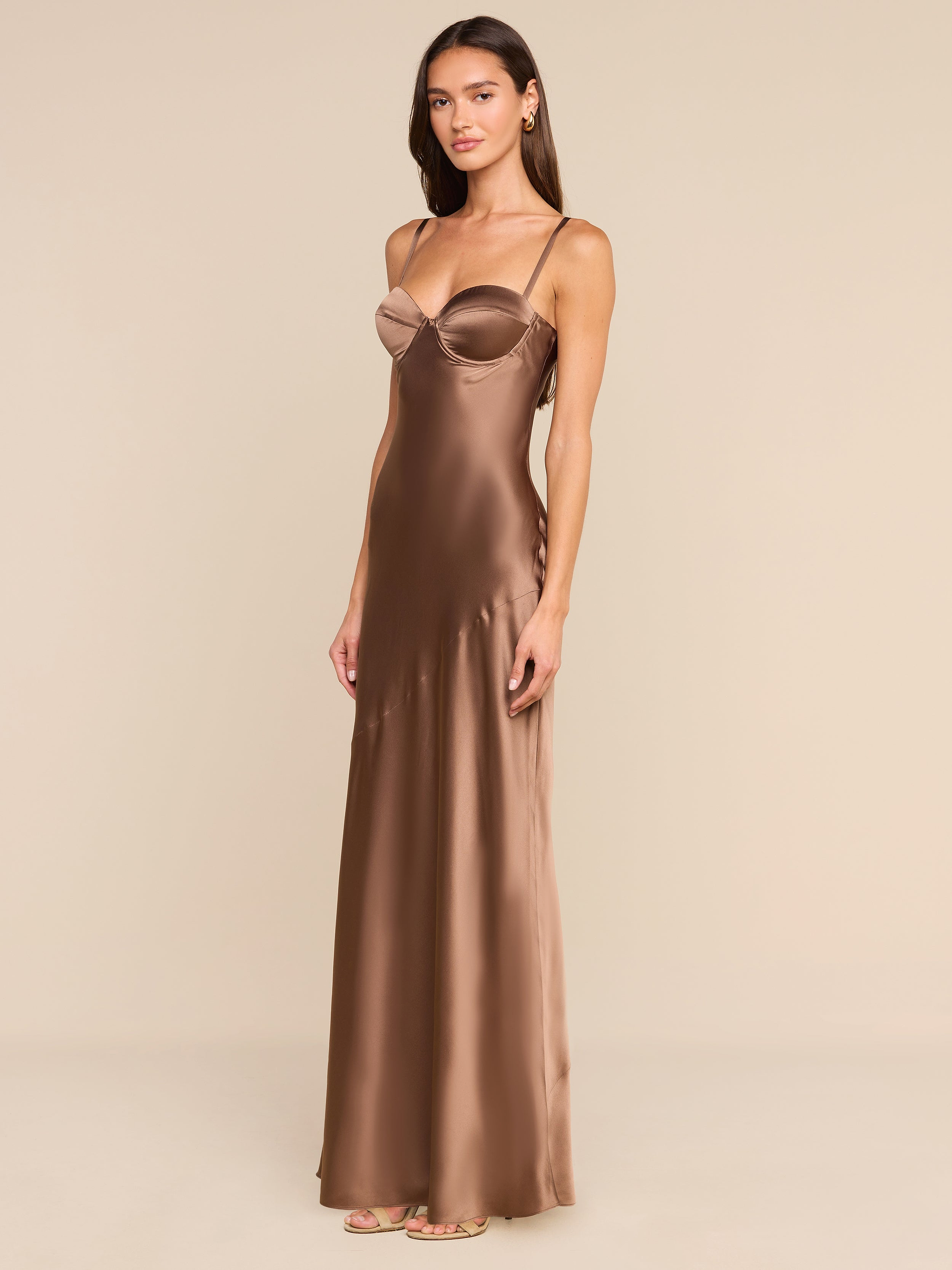 Draped in the Sayla Silk Gown—a long, bronze satin dress with delicate thin straps—the woman exudes vintage glamour as she poses against a plain white background, gazing at the camera with one hand resting by her side.