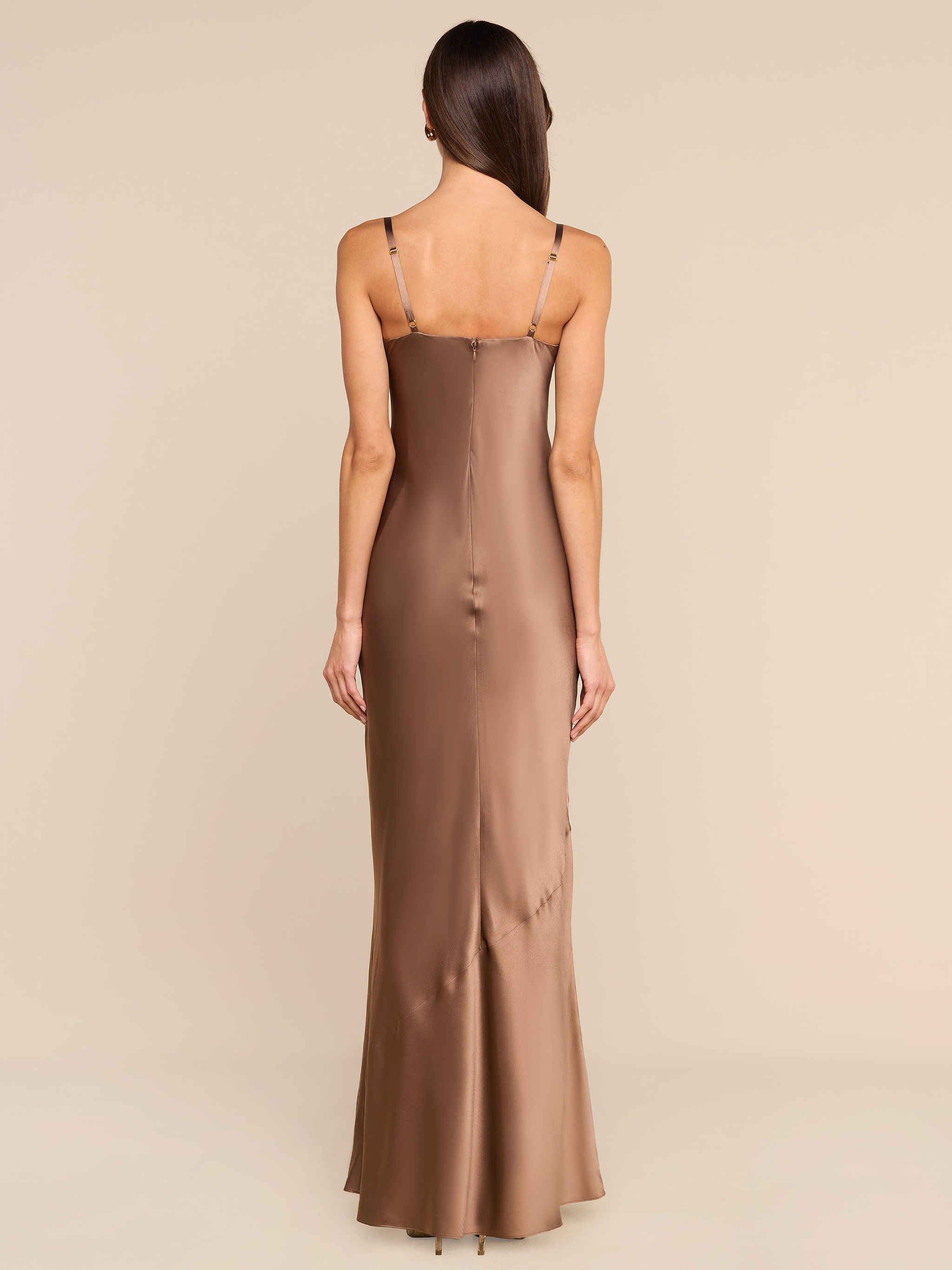 A woman with long brown hair stands facing away, wearing the Sayla Silk Gown—a sleeveless, floor-length bronze satin dress with thin straps that exudes vintage glamour—set against a plain white background.