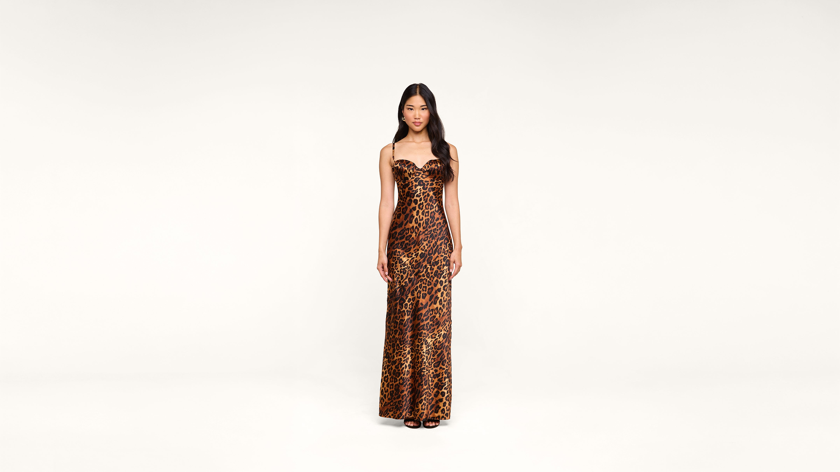 A woman stands facing forward, wearing a long, form-fitting leopard print dress with thin straps and black open-toed shoes, against a plain white background.
