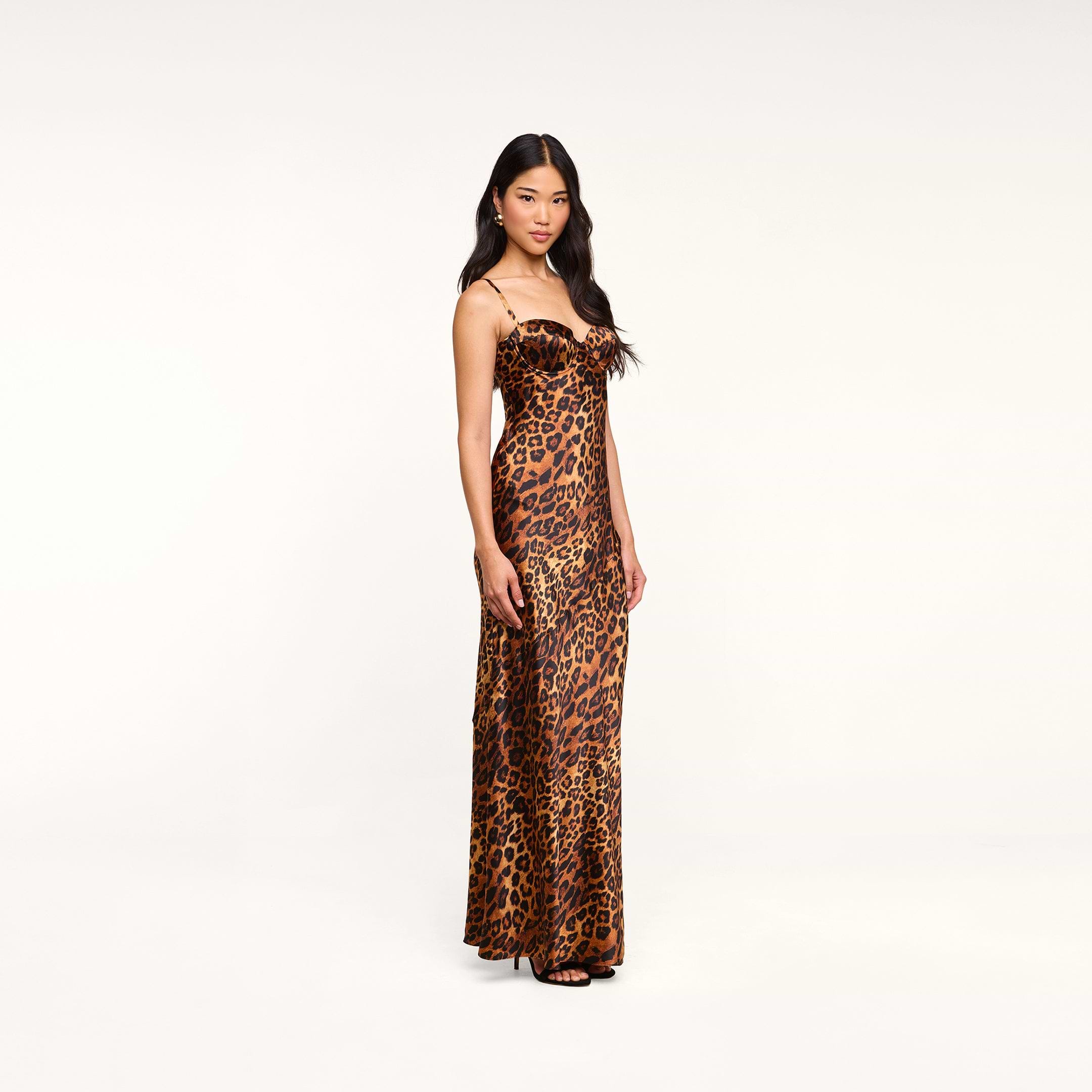 A woman stands in the Sayla Silk Gown—a full-length, sleeveless dress with thin straps and a leopard print—posing against a plain white background, creating an event-ready look.