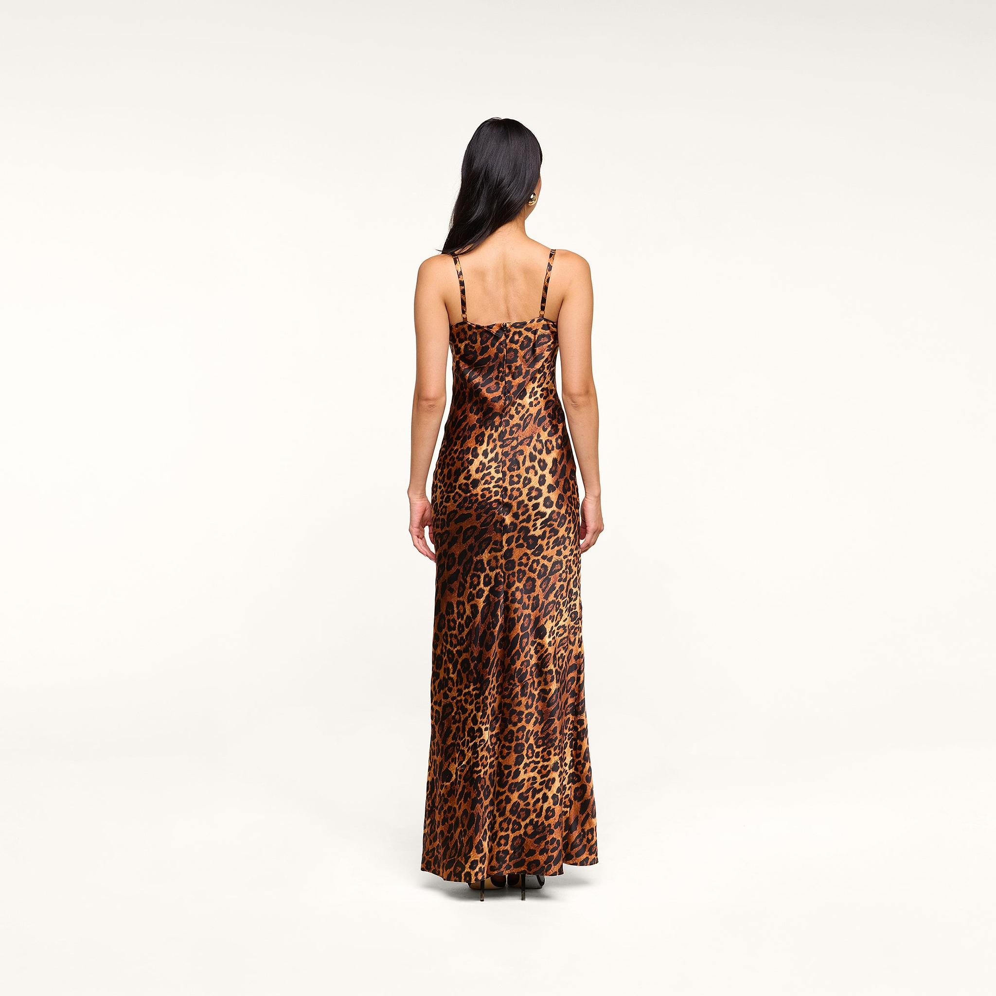 A woman with long dark hair, seen from behind, wears the Sayla Silk Gown—a floor-length, sleeveless dress in a leopard print with thin straps—standing against a plain light background.