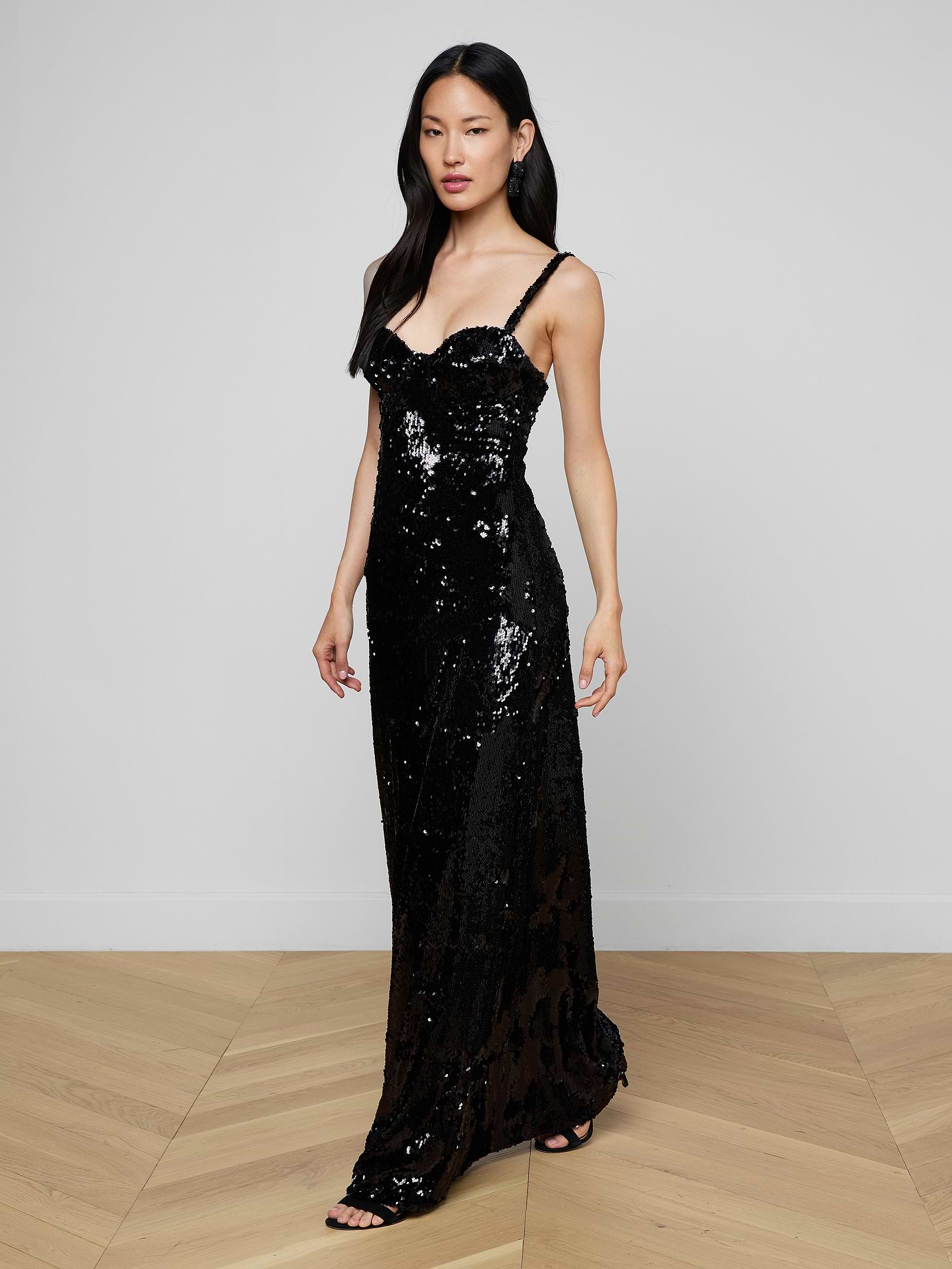 A woman wearing the Sayla Sequin Gown—a long, black sequin dress with thin straps that exudes vintage glamour—stands against a plain light background.