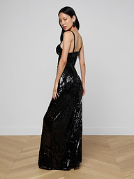 Draped in the Sayla Sequin Gown—a long, sleeveless black sequin dress—a woman stands against a plain light background, channeling vintage glamour as she gazes over her shoulder with one hand resting by her side.