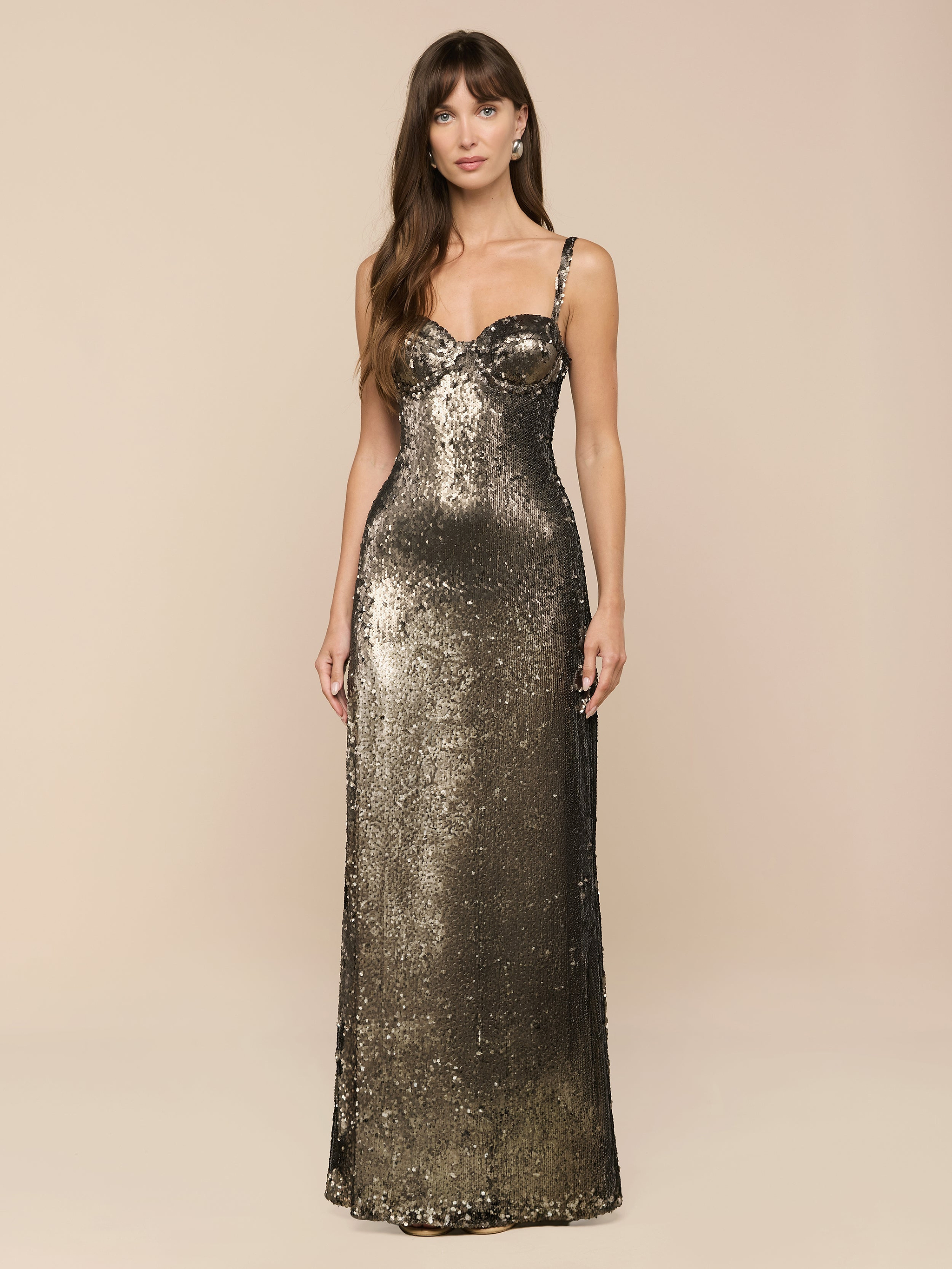 A woman stands facing forward against a plain white background, radiating vintage glamour in the Sayla Sequin Gown—a long, fitted dress with thin straps and dazzling olive green sequins.