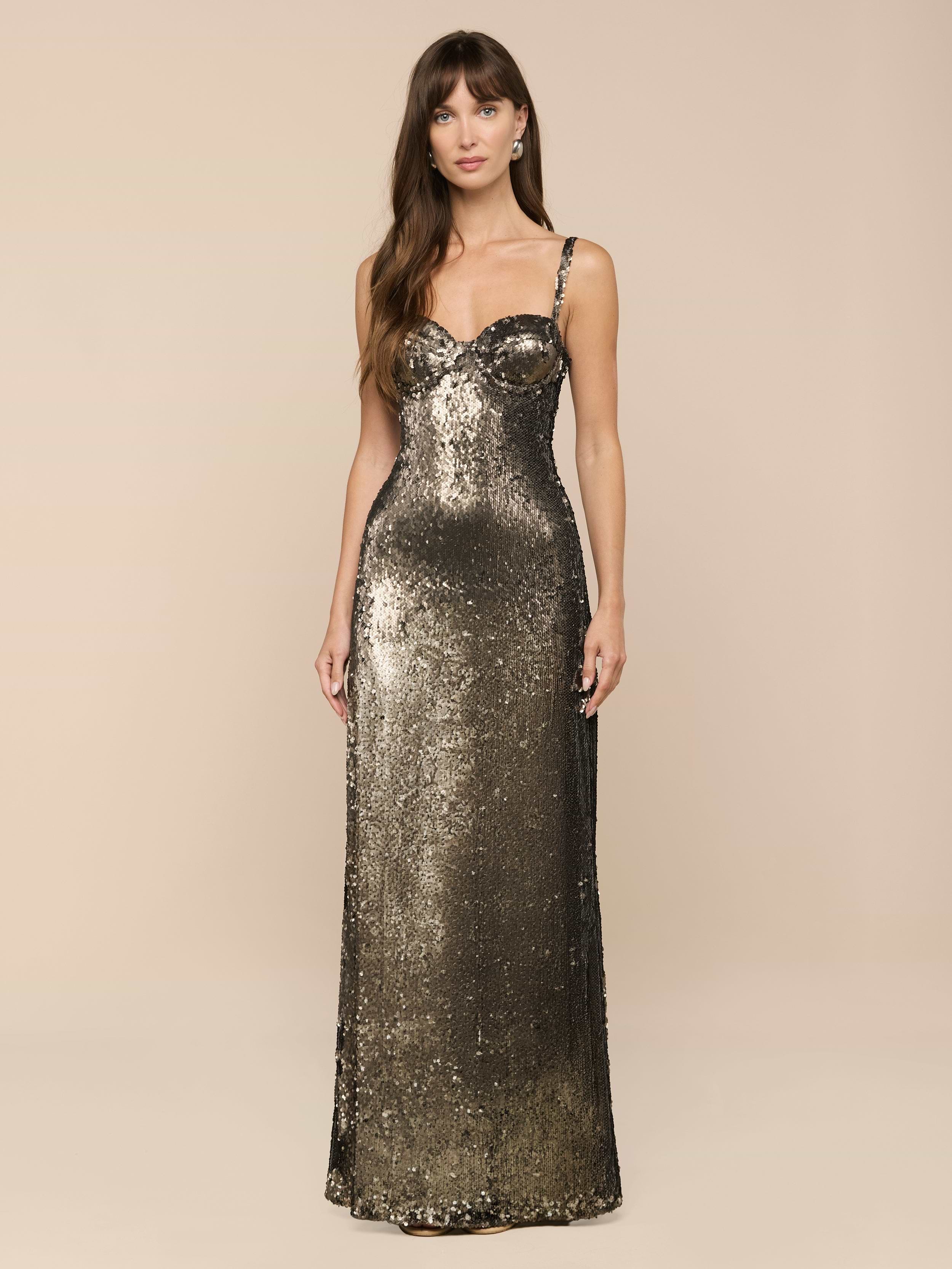 A woman stands facing forward against a plain white background, radiating vintage glamour in the Sayla Sequin Gown—a long, fitted dress with thin straps and dazzling olive green sequins.