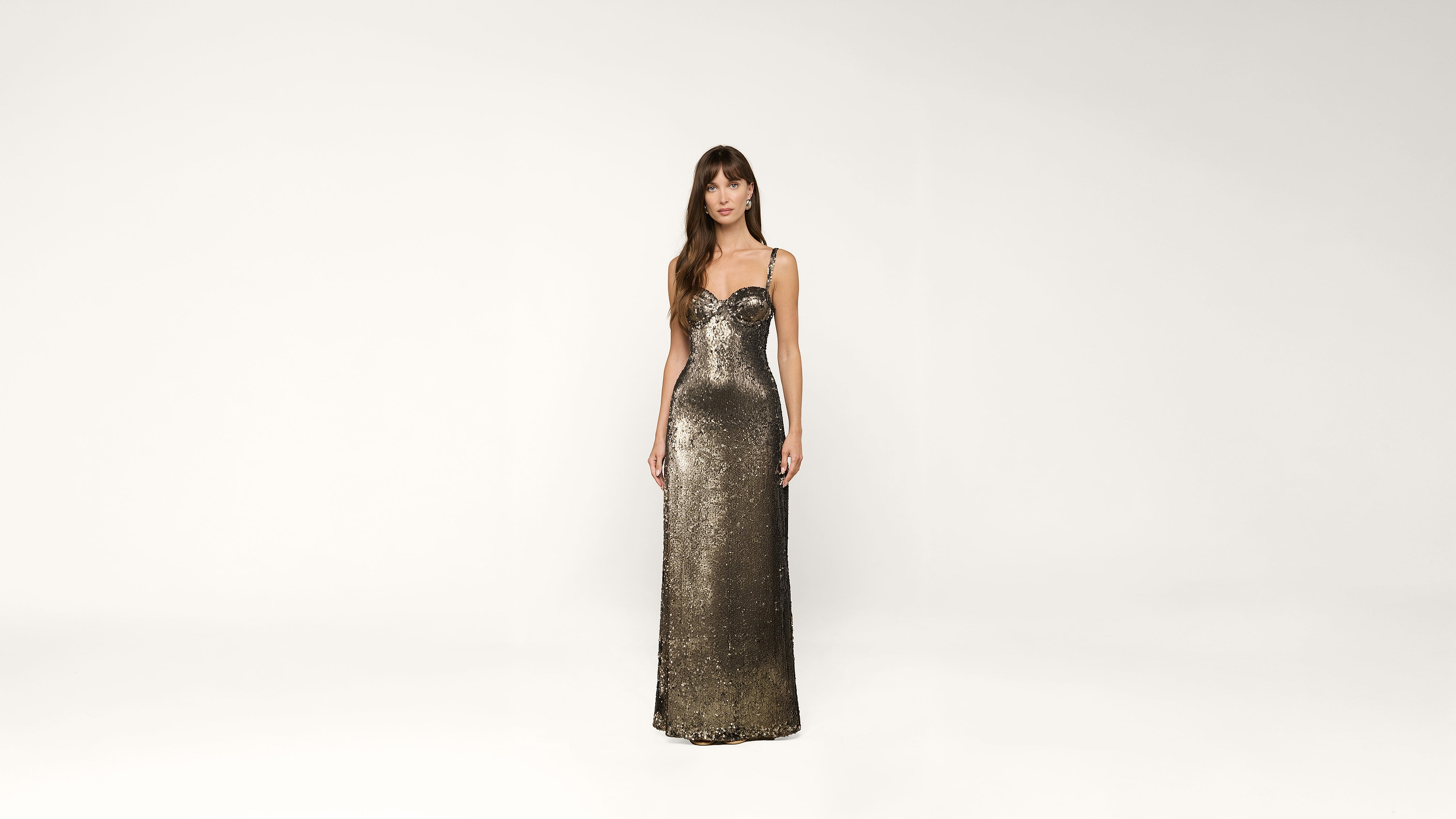 A woman in a metallic, floor-length, fitted sequin gown stands against a plain white background, looking forward with relaxed arms at her sides.
